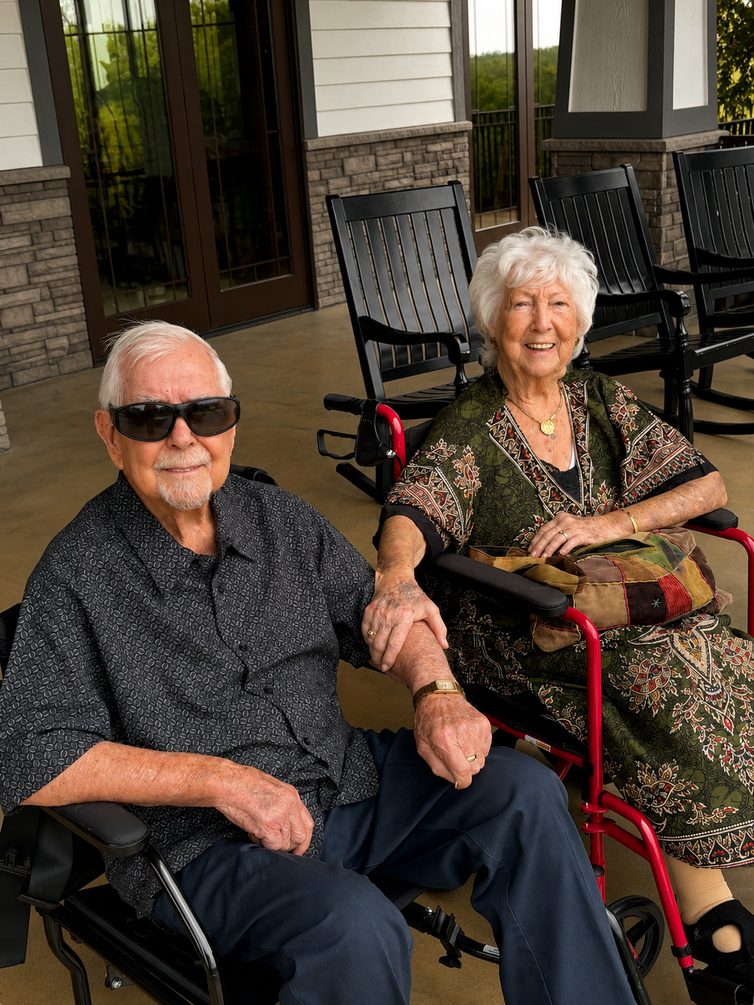 An elderly man and woman sitting outdoors on a porch. The man wears sunglasses and a patterned shirt, while the woman is in a patterned dress and sitting in a wheelchair. They are holding hands and smiling, with chairs and a brick and siding house in the background.