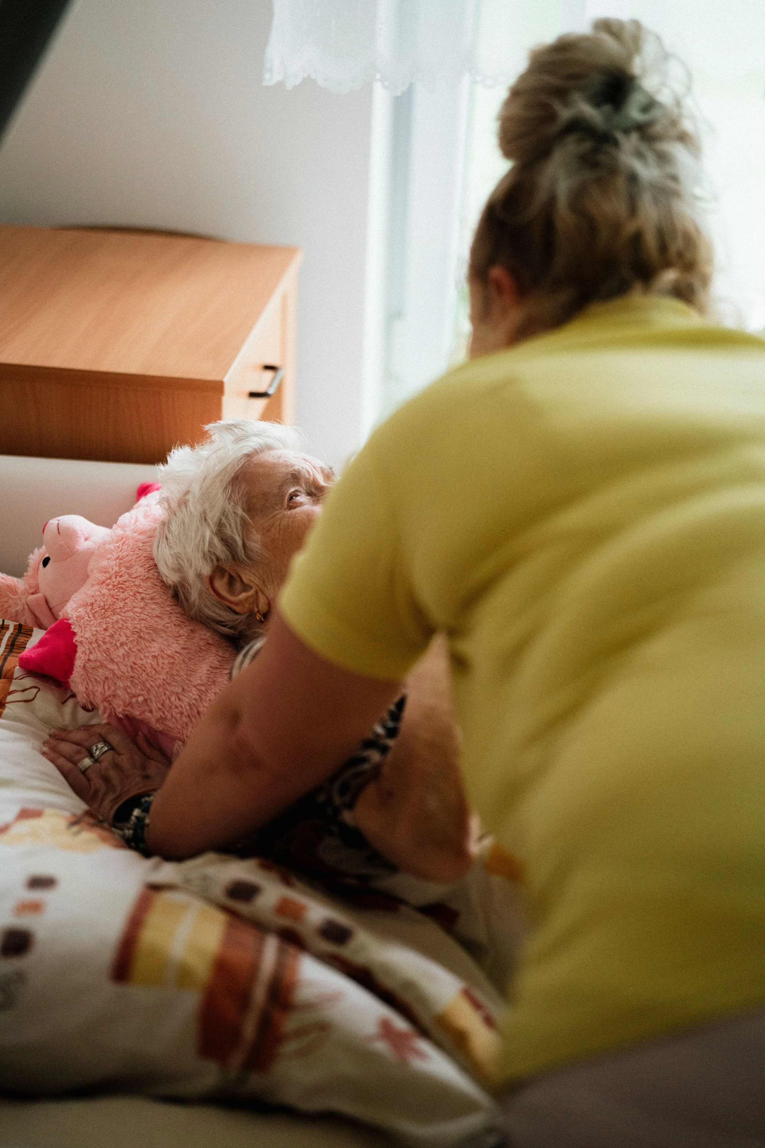 An elderly woman lying in bed, wearing a pink fleece blanket, looking up at a person standing next to her. The person is wearing a yellow shirt with their back turned to the camera. The room has a window with lace curtains, and a wooden bedside table is visible.