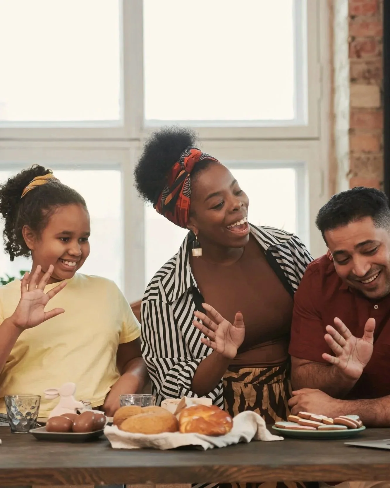 A group of three people, two women and a man, are smiling and celebrating around a table with Easter-themed treats, including chocolate eggs, bread, and cookies, in a bright room with large windows.