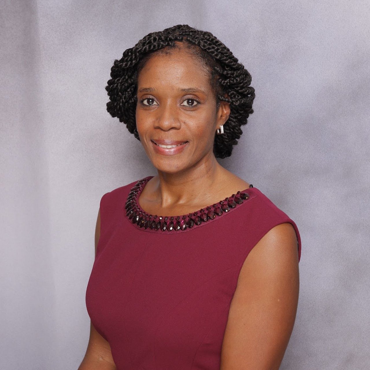 A woman with short, curly black hair wearing a sleeveless burgundy dress with decorative embellishments around the neckline, standing against a light gray background, smiling at the camera.
