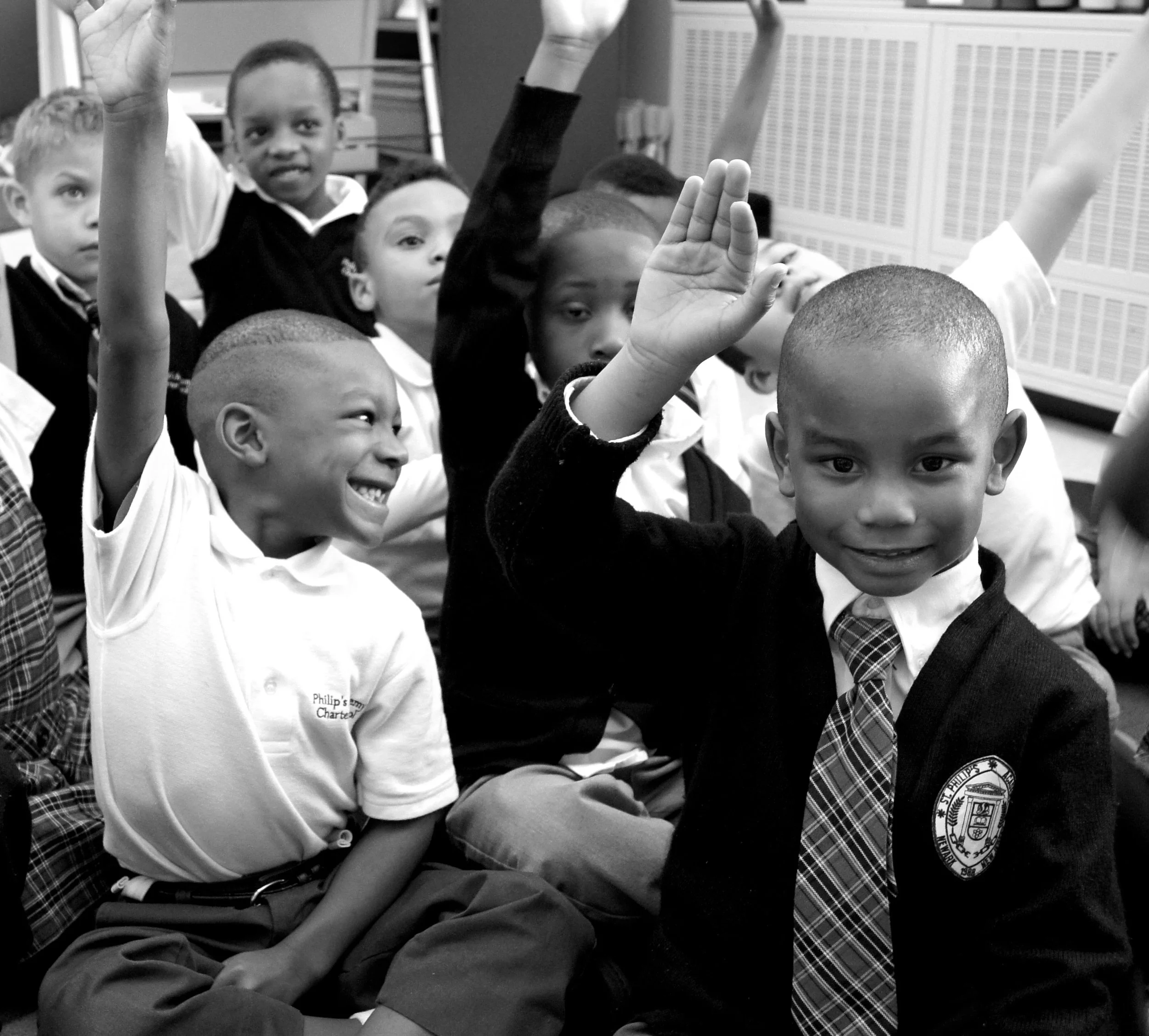 A group of young school children, mostly boys, sitting and raising their hands during a classroom activity. Some children are smiling while others are focused.