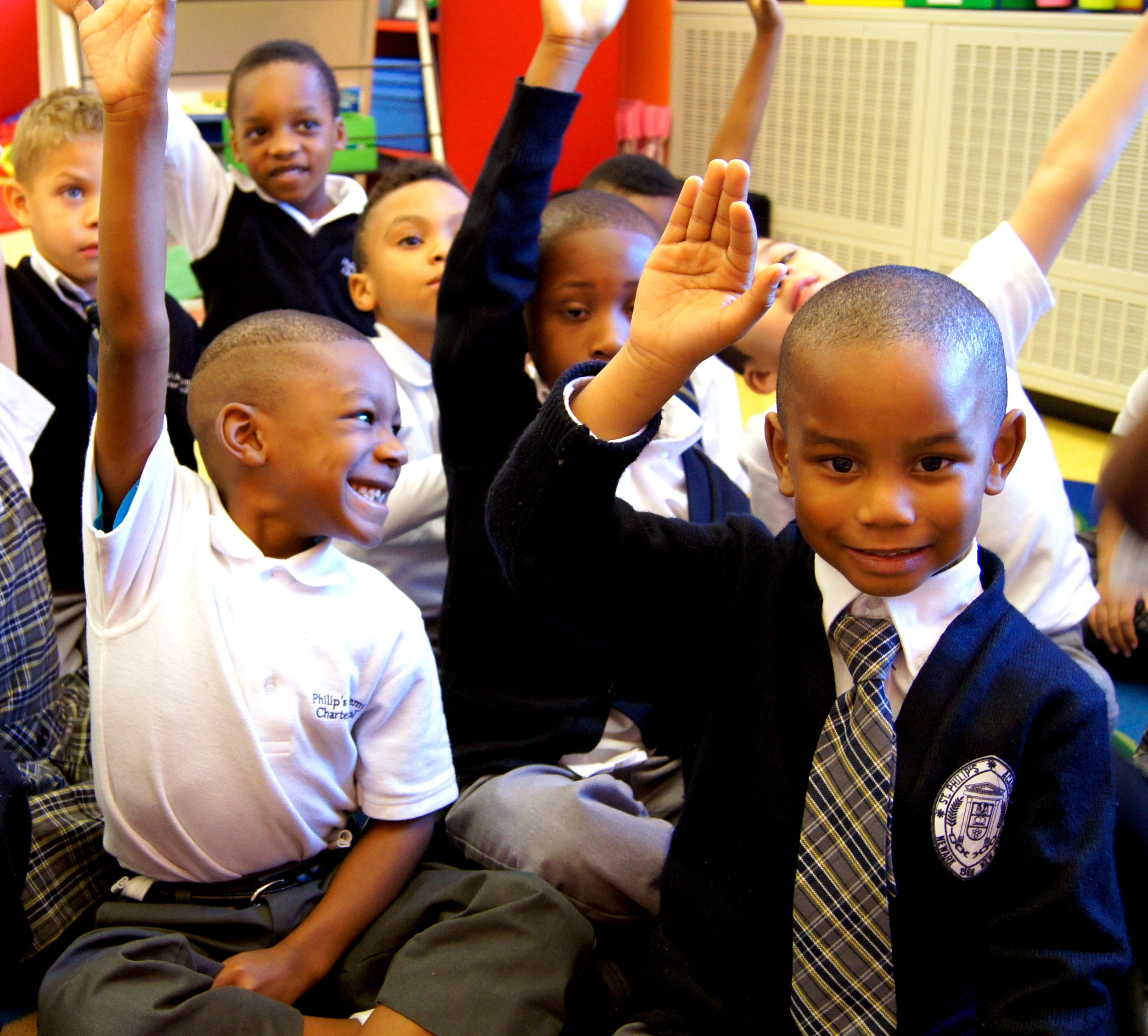 A group of schoolchildren in uniforms sitting in a classroom raising their hands for a question or answer.