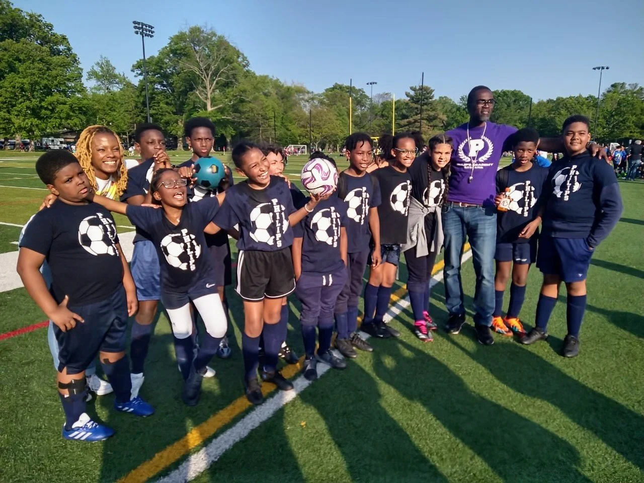 A youth soccer team with players and coaches posing on a soccer field during daytime for a group photo, all wearing matching dark blue jerseys with white soccer ball logos.