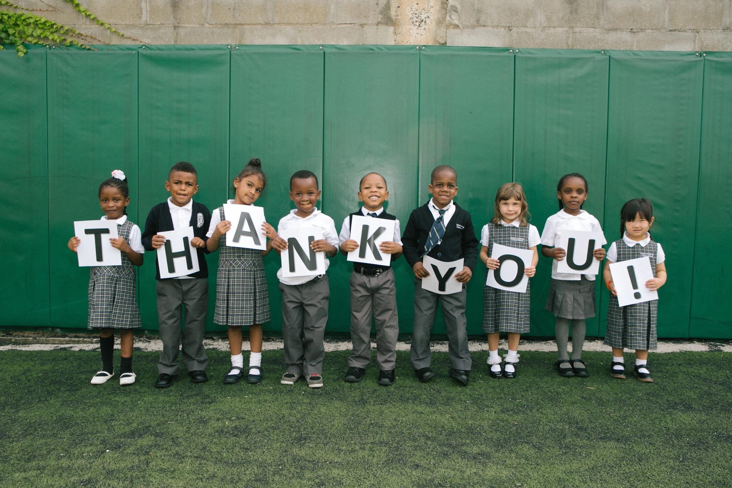 A group of nine children standing outdoors against a green background, holding up letters that spell 'THANK YOU'.