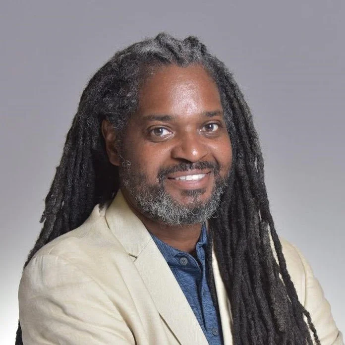 Portrait of a man with long dreadlocks and a beard, smiling, wearing a beige blazer and a blue shirt against a neutral background.