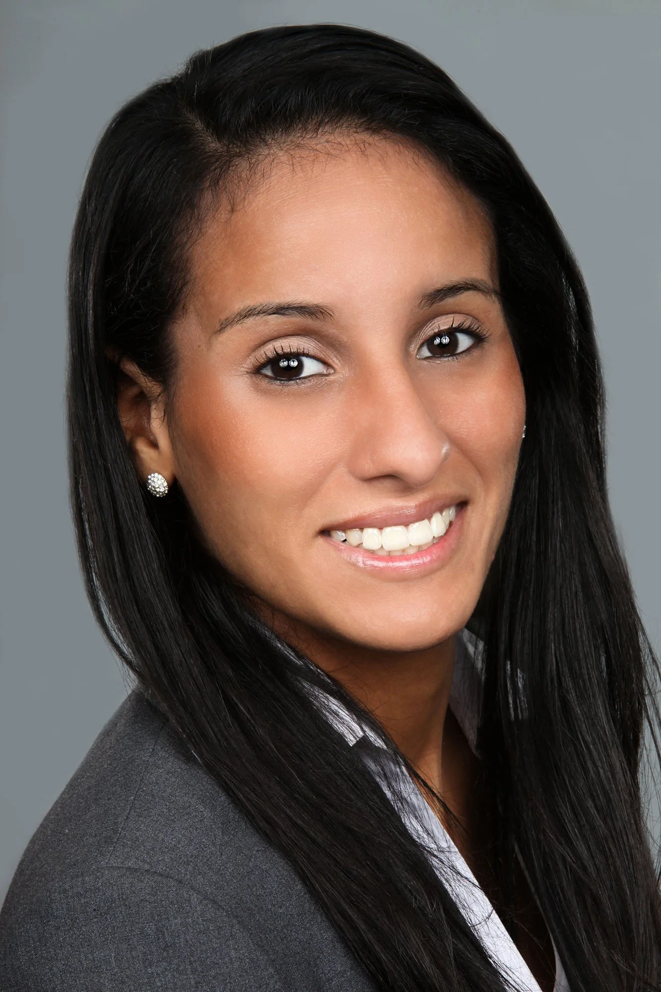 Close-up portrait of a woman with long black hair, wearing a gray blazer and white shirt, smiling against a gray background.