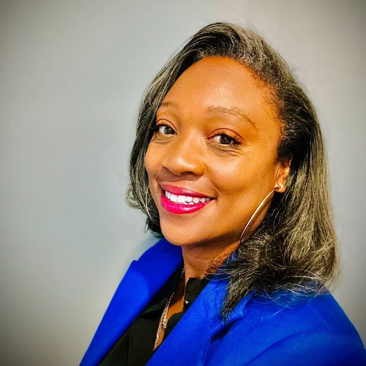 A smiling woman with shoulder-length black hair, wearing a blue blazer, earrings, and bright pink lipstick, against a gray background.