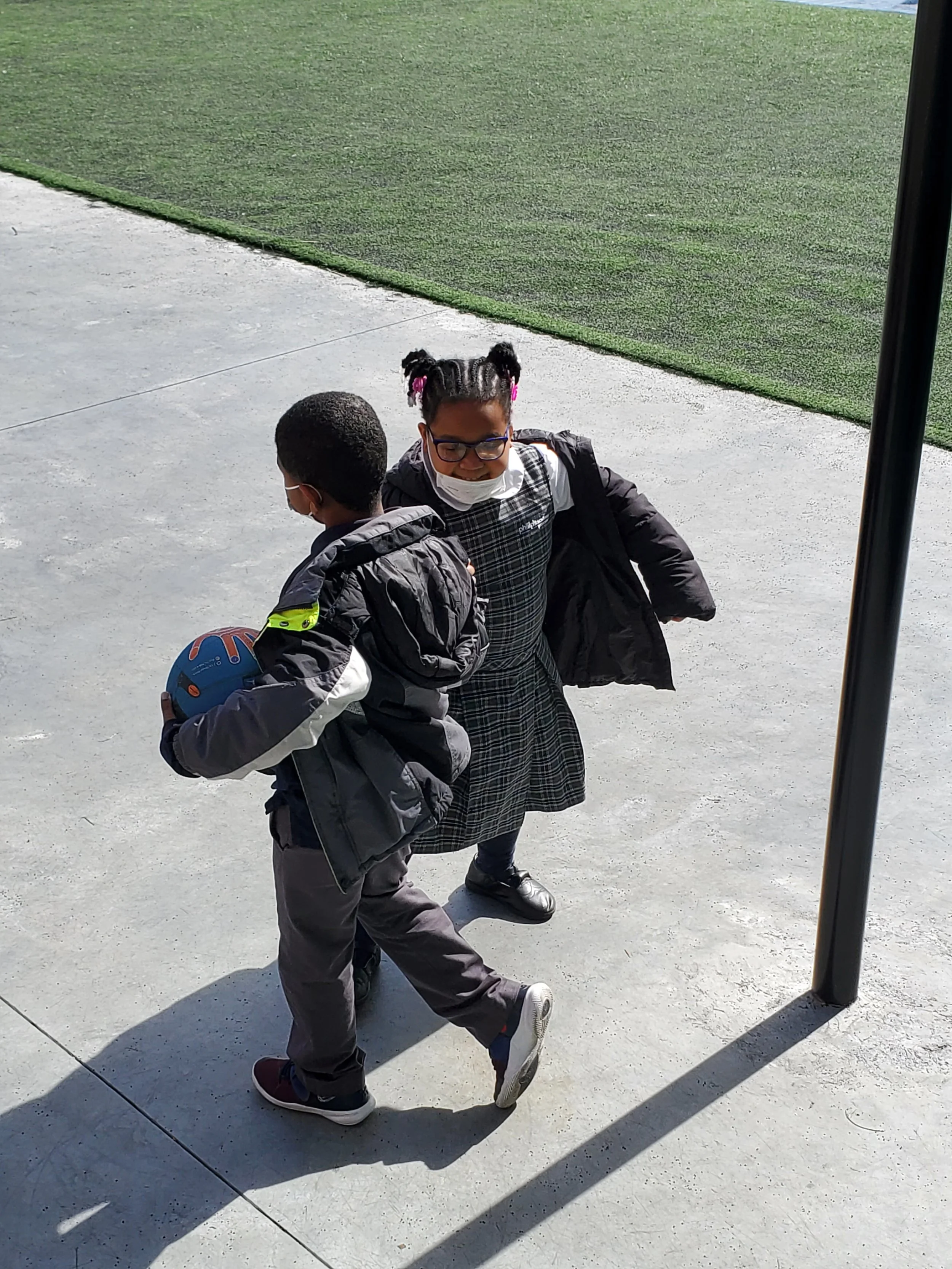 A young girl with pigtails, glasses, and a face mask, wearing a checkered dress under a black jacket, smiling as she interacts with a young boy holding a blue basketball, outside on a concrete surface with a grassy area in the background.