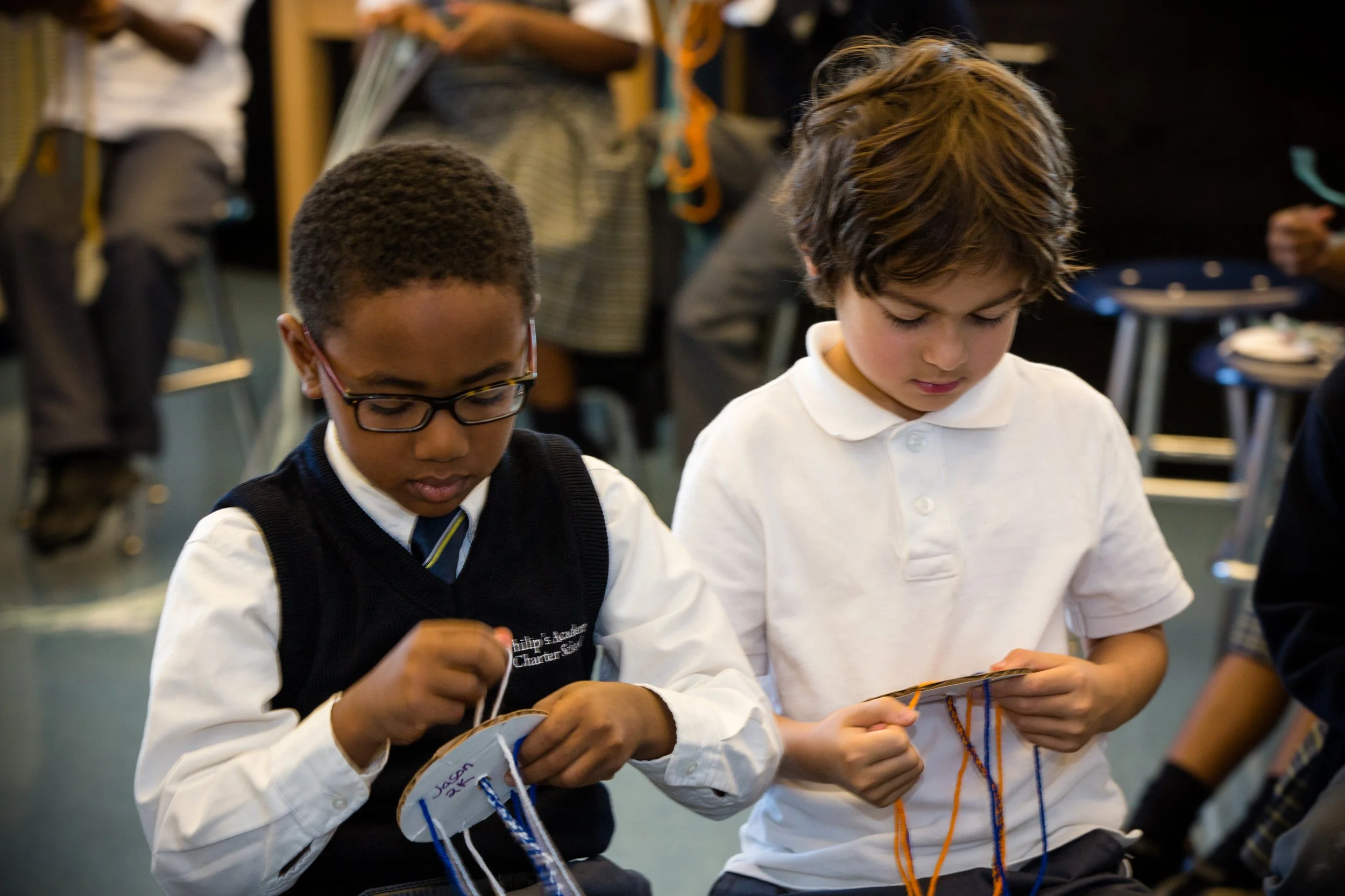 Two boys with short brown hair, one with glasses, wearing school uniforms, looking down and working on a project in a classroom.