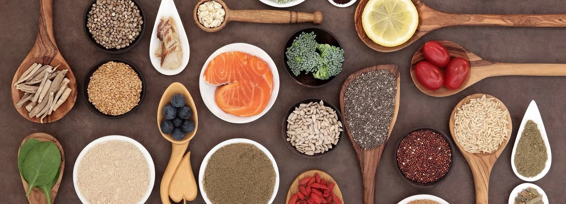 A variety of fresh ingredients and spices arranged in small bowls and on wooden spoons, including salmon, cherry tomatoes, blueberries, lemon slices, broccoli, assorted seeds, grains, herbs, and dried goji berries.