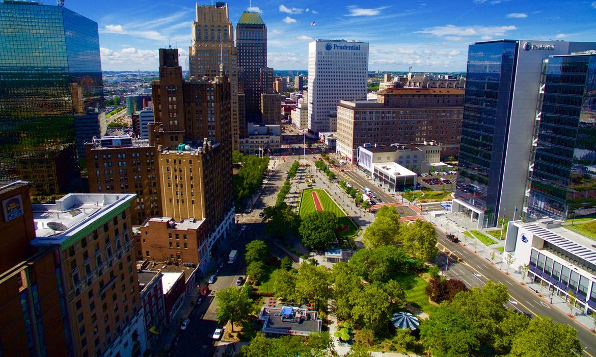 A cityscape with tall office buildings, a park with trees, and a blue sky with some clouds.