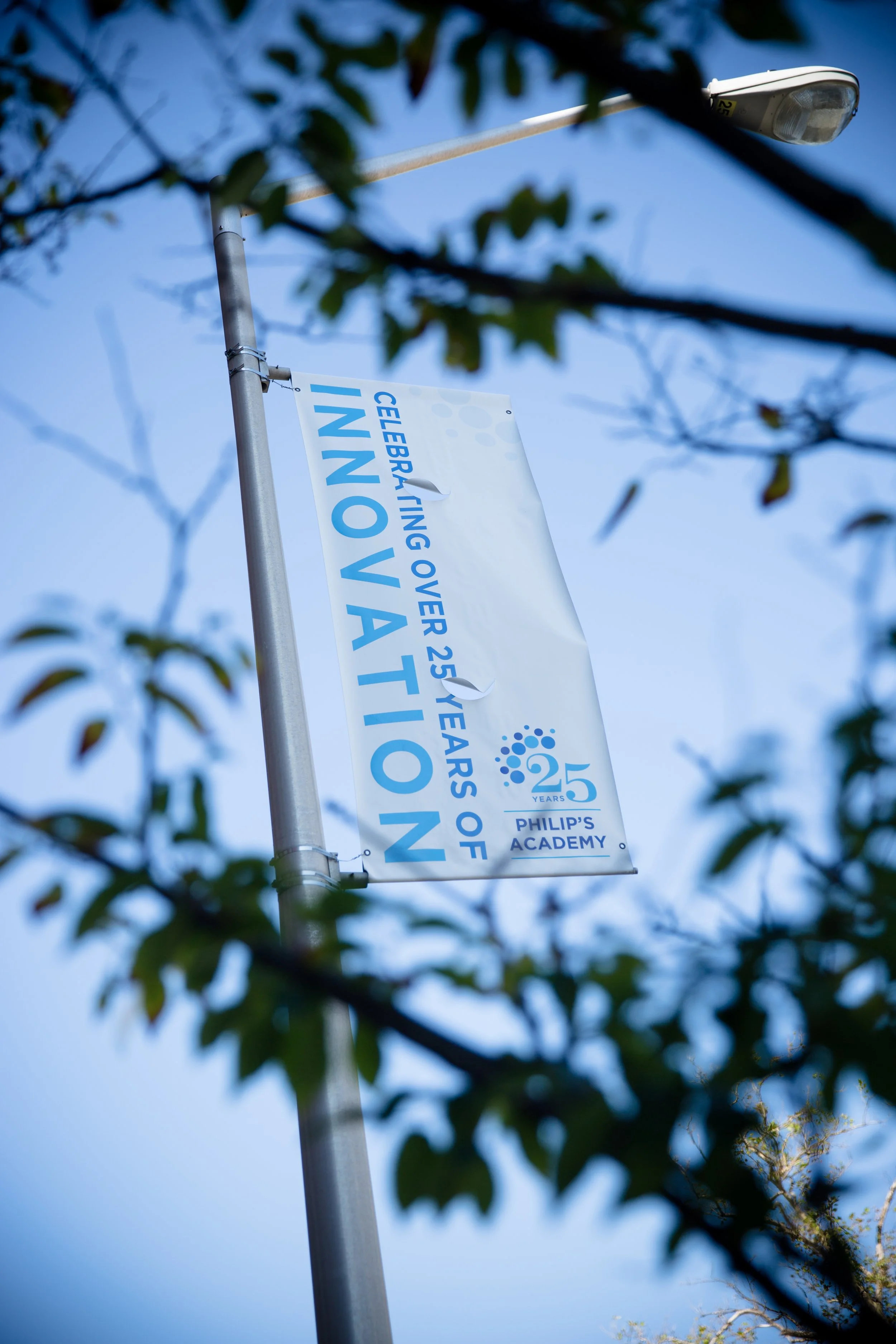 A street pole with a banner celebrating the 25th anniversary of Philip's Academy, partly obscured by leaves and branches, with a clear blue sky in the background.