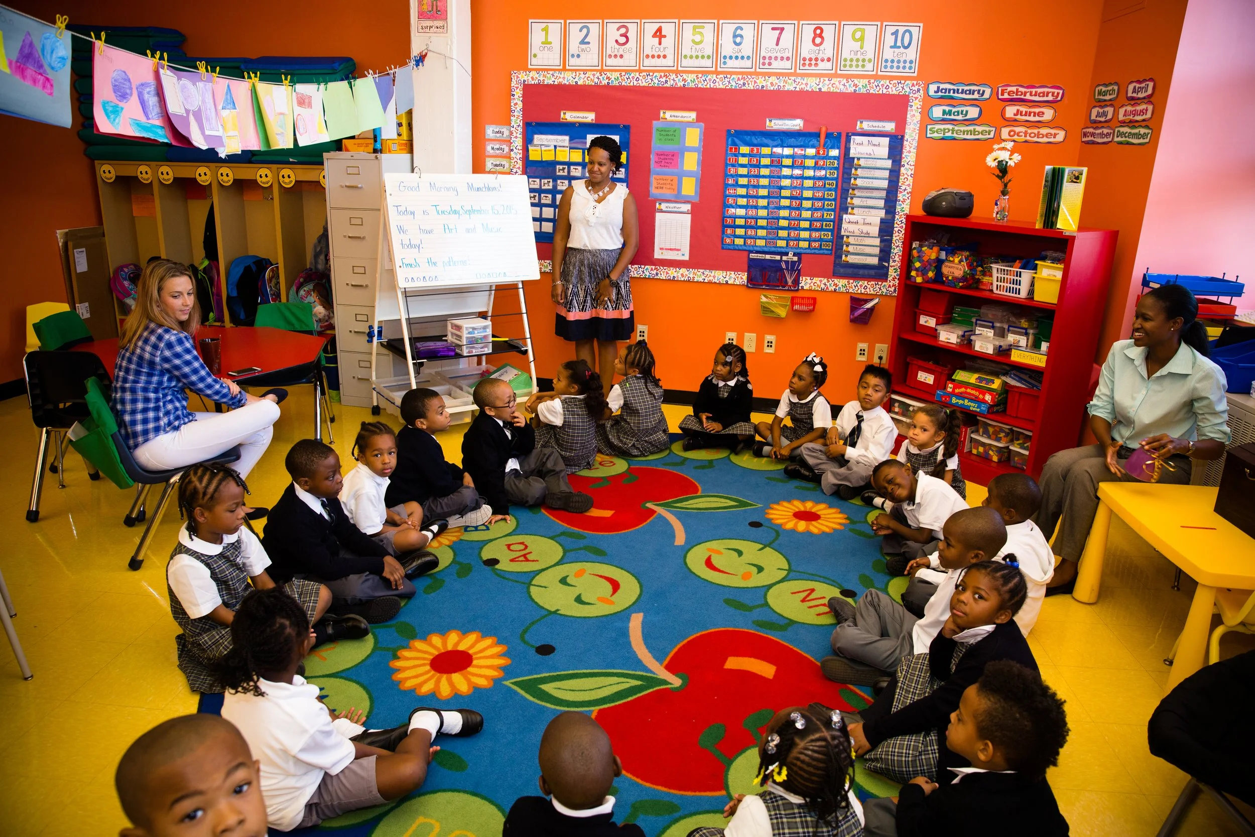 Kindergarten classroom with young students sitting on a colorful carpet in a semi-circle, teachers, and educational posters on the orange and purple walls.