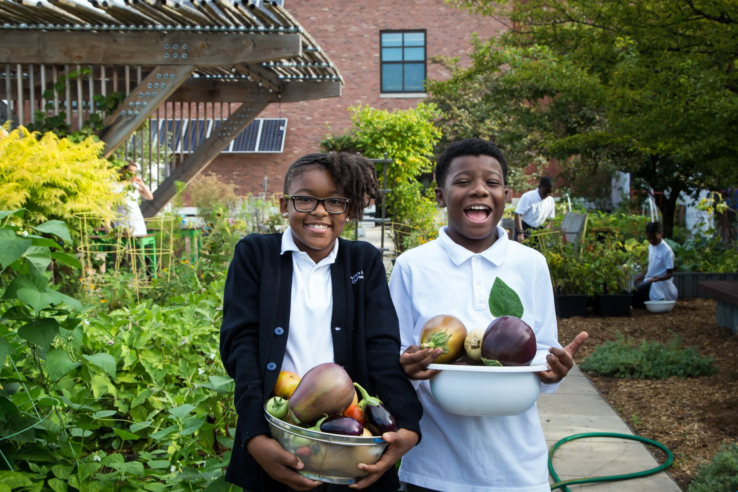 Two children in school uniforms standing in a garden, holding a bowl and a basket of freshly picked vegetables, smiling.