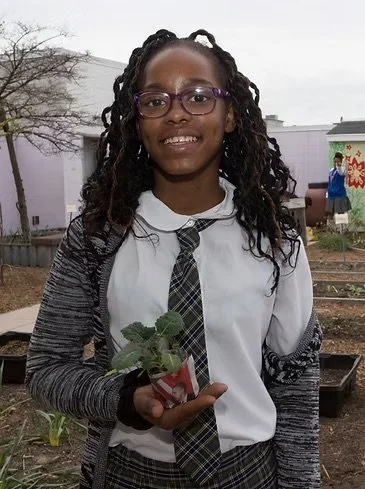 Young girl in school uniform holding a small plant in a garden