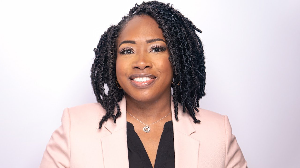 A woman with dark curly hair, wearing a light pink blazer over a black top, smiling against a plain white background.