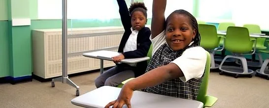 Two children in a classroom, one smiling and raising her hand, the other sitting at a desk with her hand on her chin.