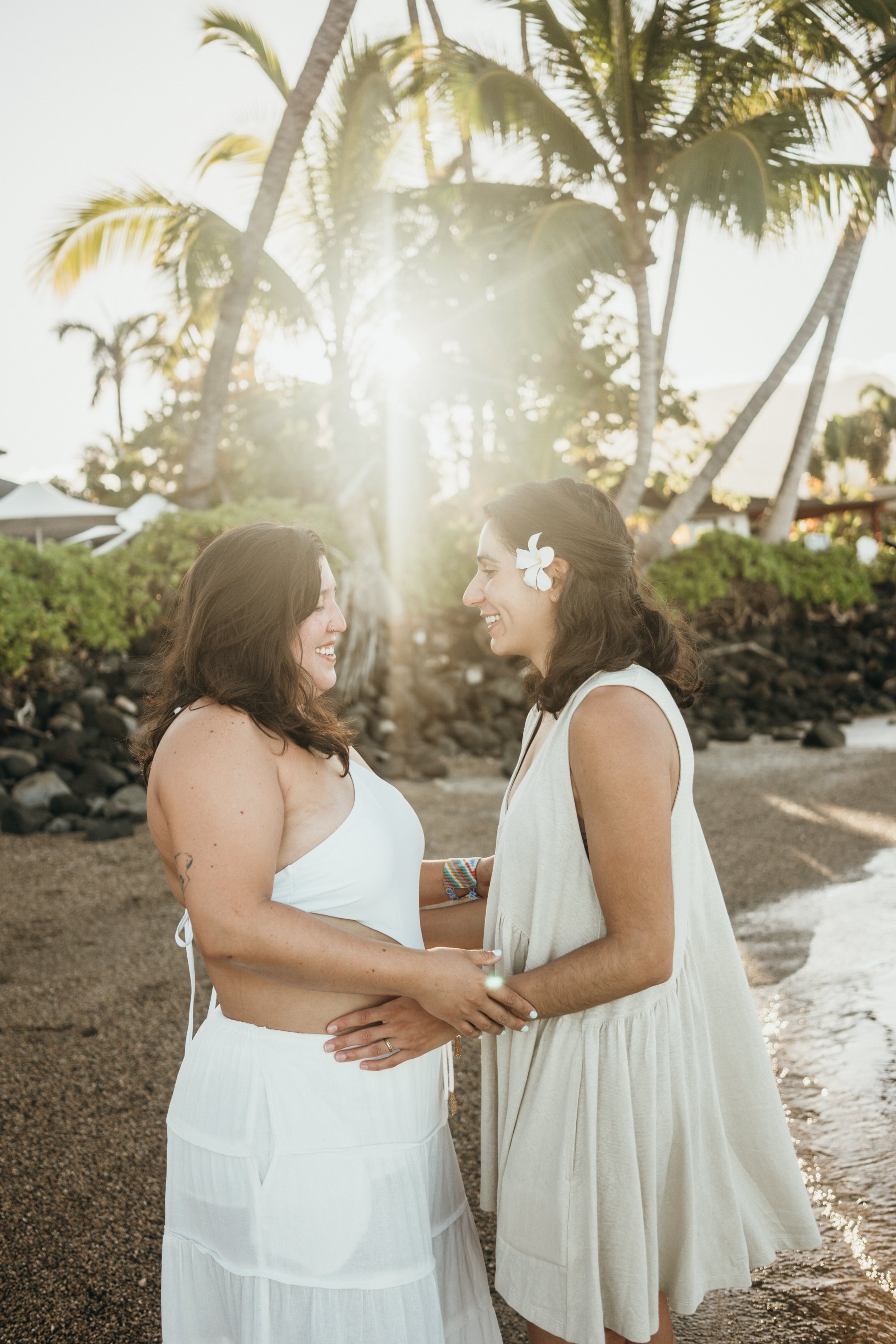 A same-sex couple shares an emotional post-proposal moment on a tropical beach at sunrise. The lighting is soft and golden, enhancing the intimacy of the scene. The composition focuses on natural interaction and storytelling rather than staging. This