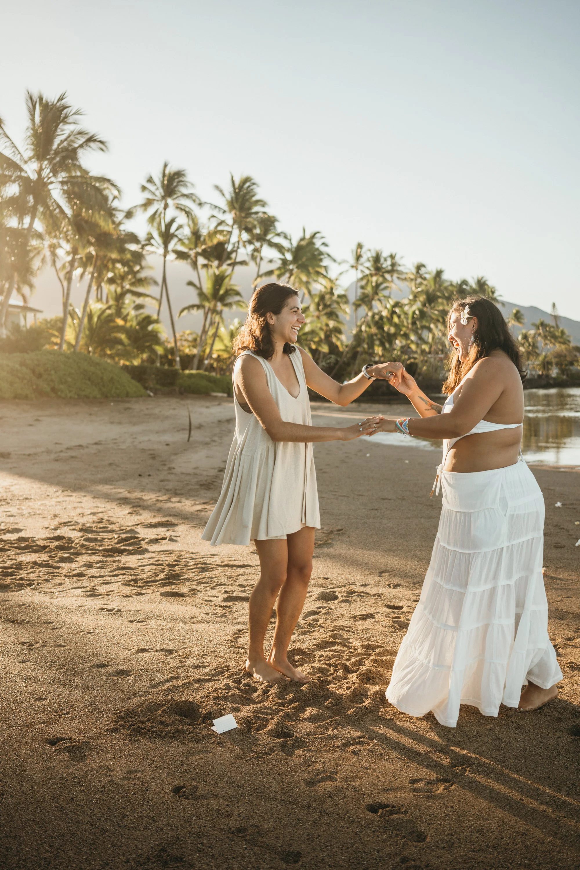 This is a same-sex engagement proposal captured on a beach at sunrise. The soft light creates a warm and emotional atmosphere as the couple stands near the shoreline. The moment reflects authentic connection and surprise immediately after the proposa