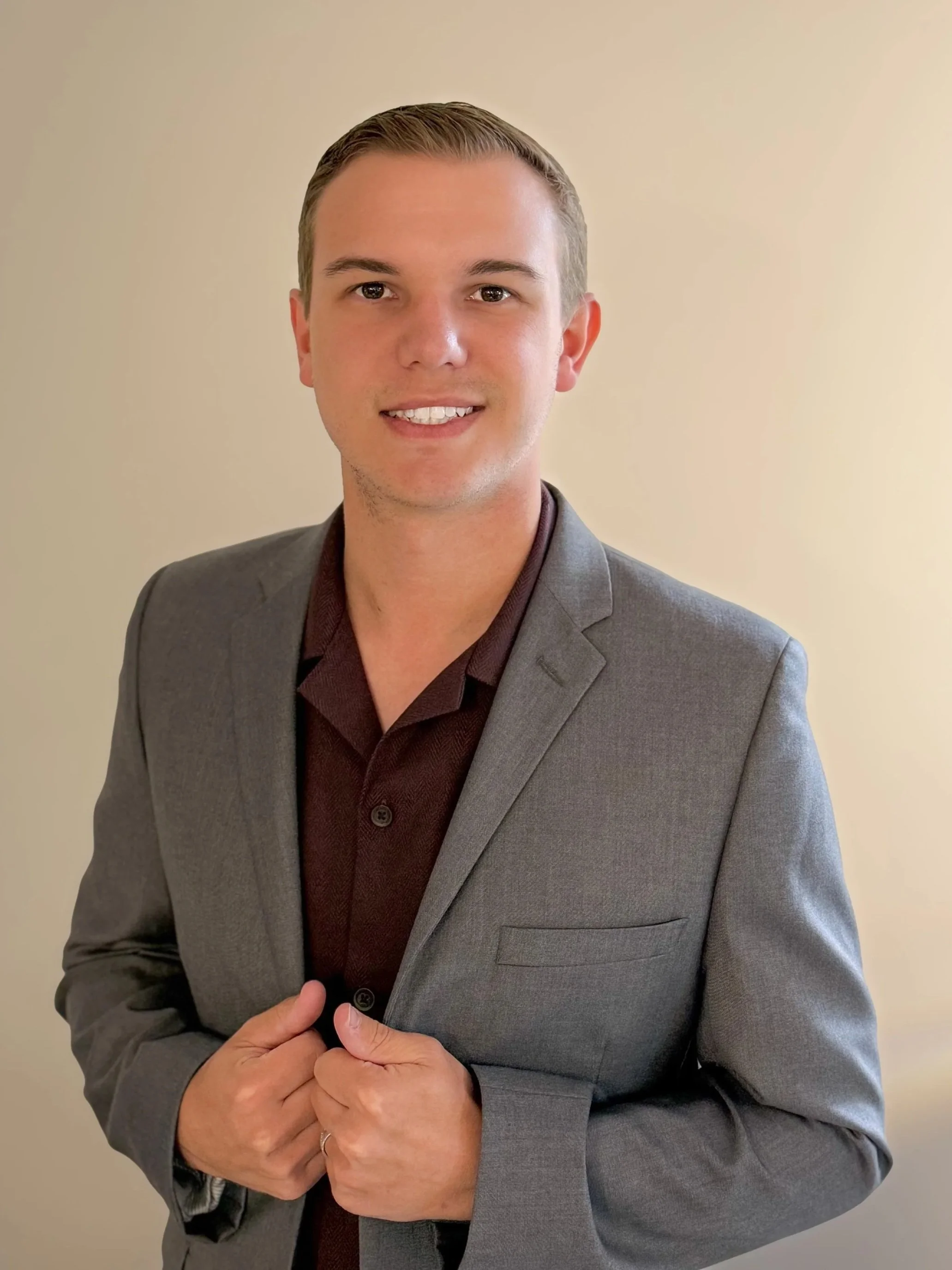 A man wearing a gray suit and maroon shirt, smiling, with hands clasped, standing against a plain beige background.