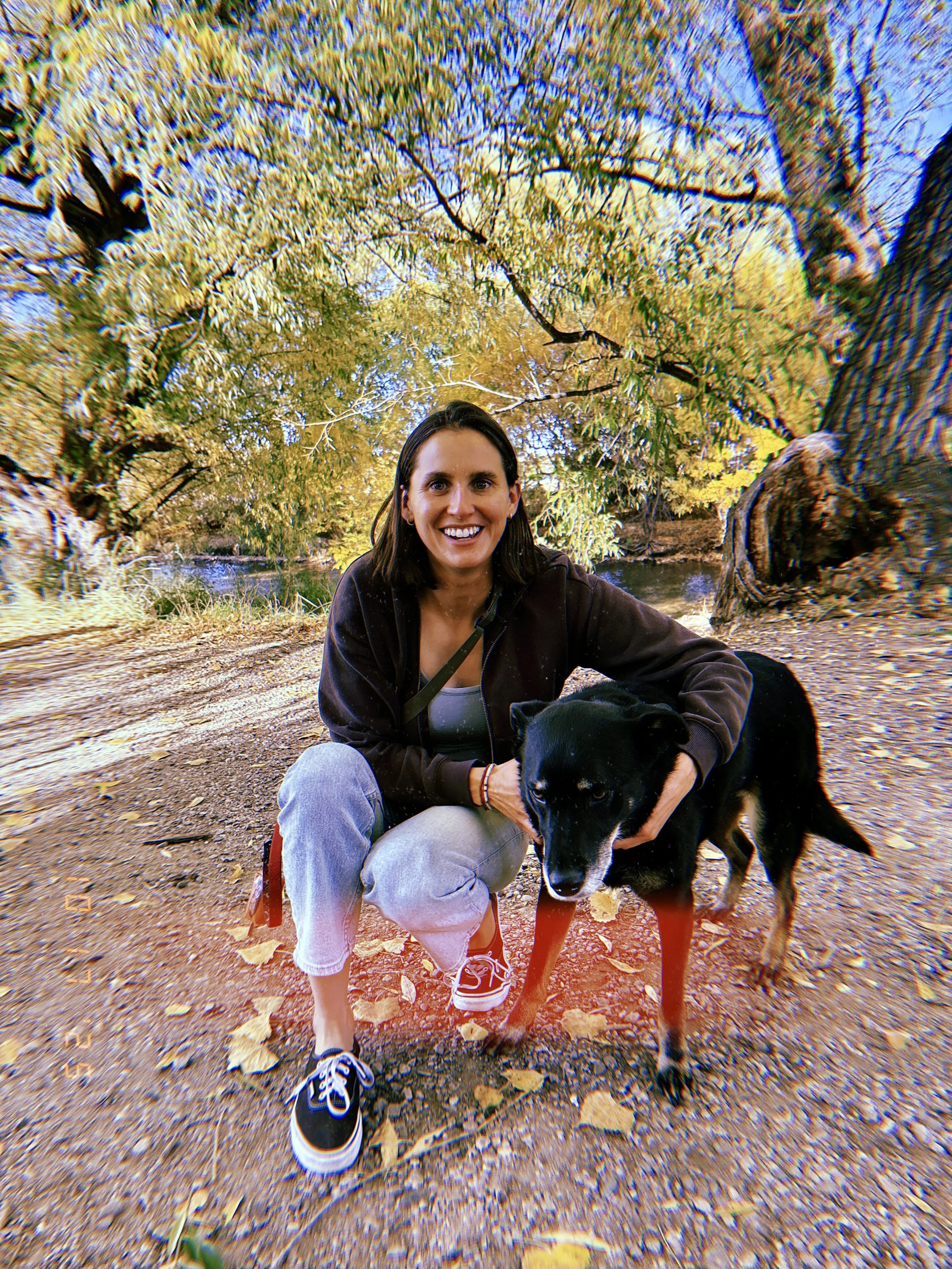 A woman in a black hoodie and gray pants kneels on a trail in a park, smiling and holding a black and white dog, with trees and fallen leaves in the background.