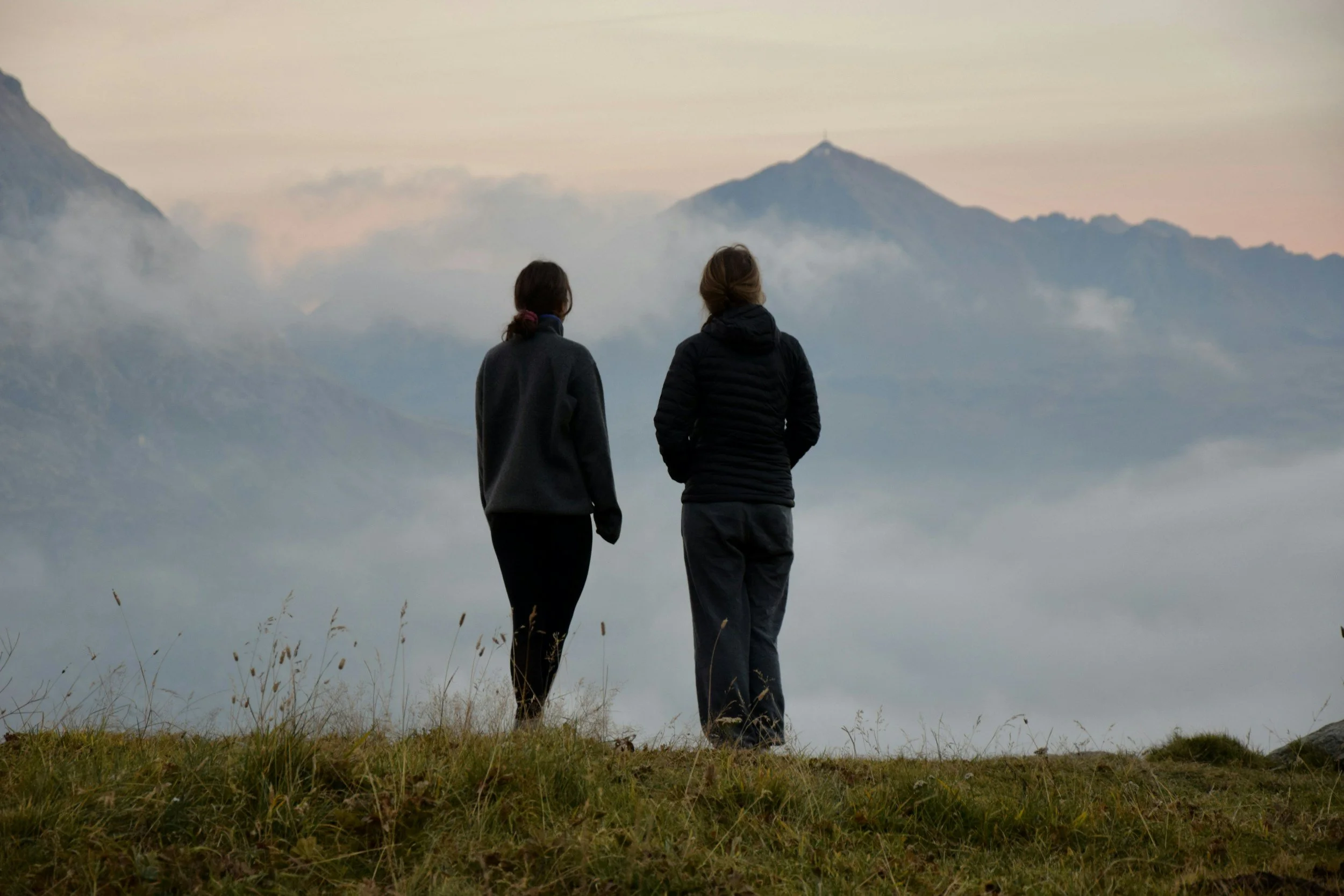 Two people standing on a grassy hilltop overlooking a mountain landscape with clouds and distant mountains in the background during dawn or dusk.
