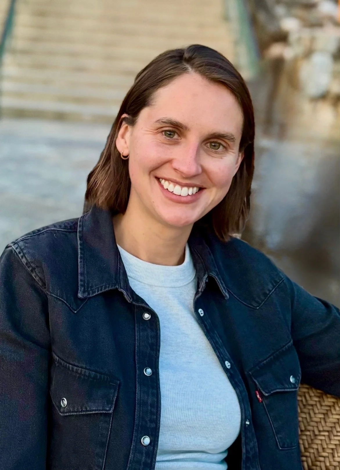 A woman with brown hair smiling outdoors near water and a bridge, wearing a dark denim jacket over a gray t-shirt.