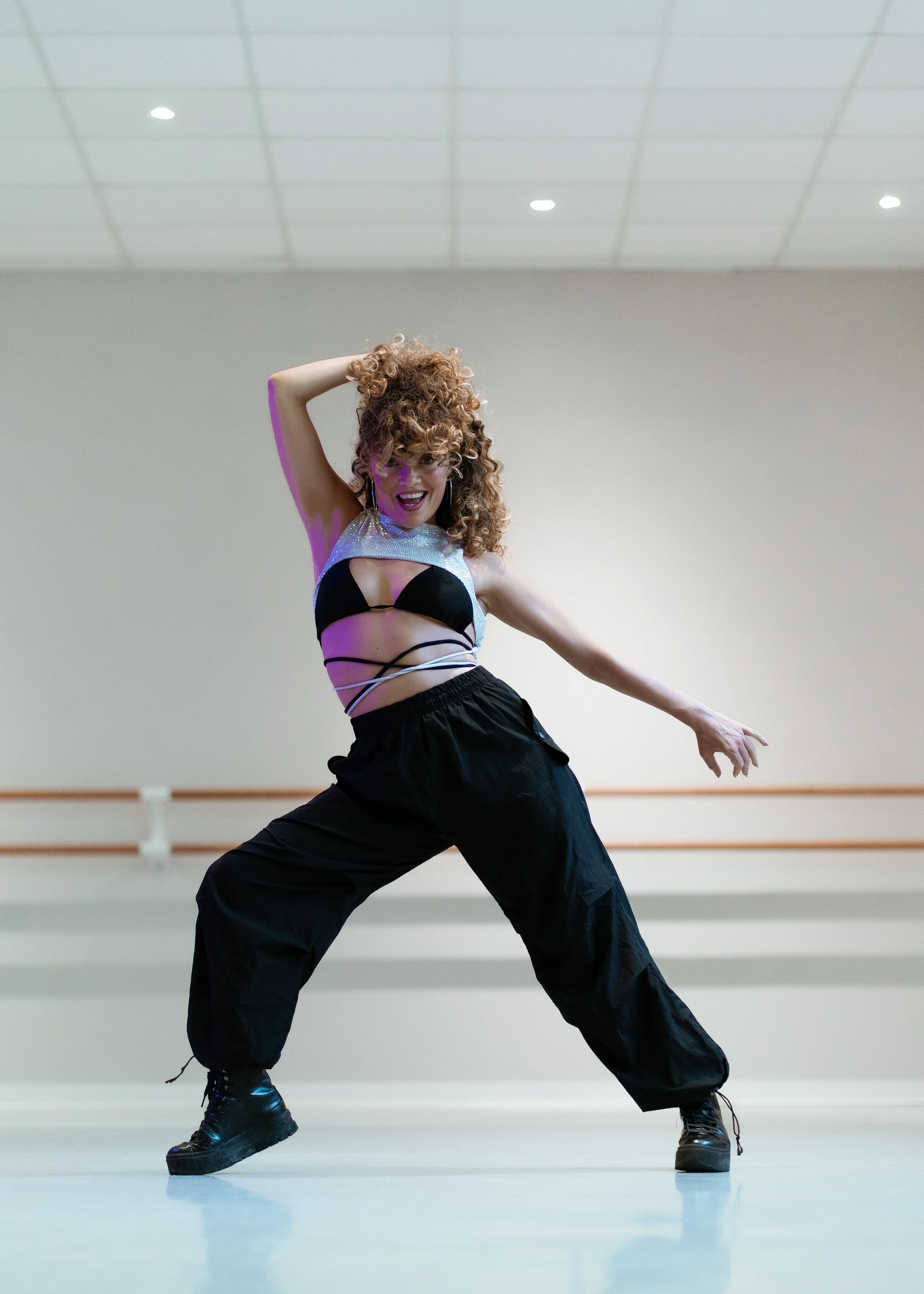 Une femme danse dans un studio avec un sol clair et un mur gris, portant un haut noir, un débardeur argenté, un pantalon ample noir, et des bottes noires. Elle a les cheveux bouclés et roux, et affiche une expression joyeuse.