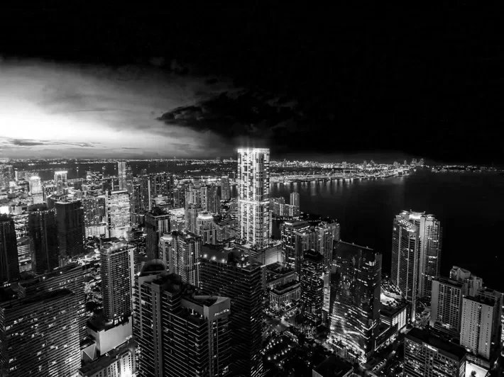 Black and white aerial view of a city skyline at night with illuminated skyscrapers and water in the background.