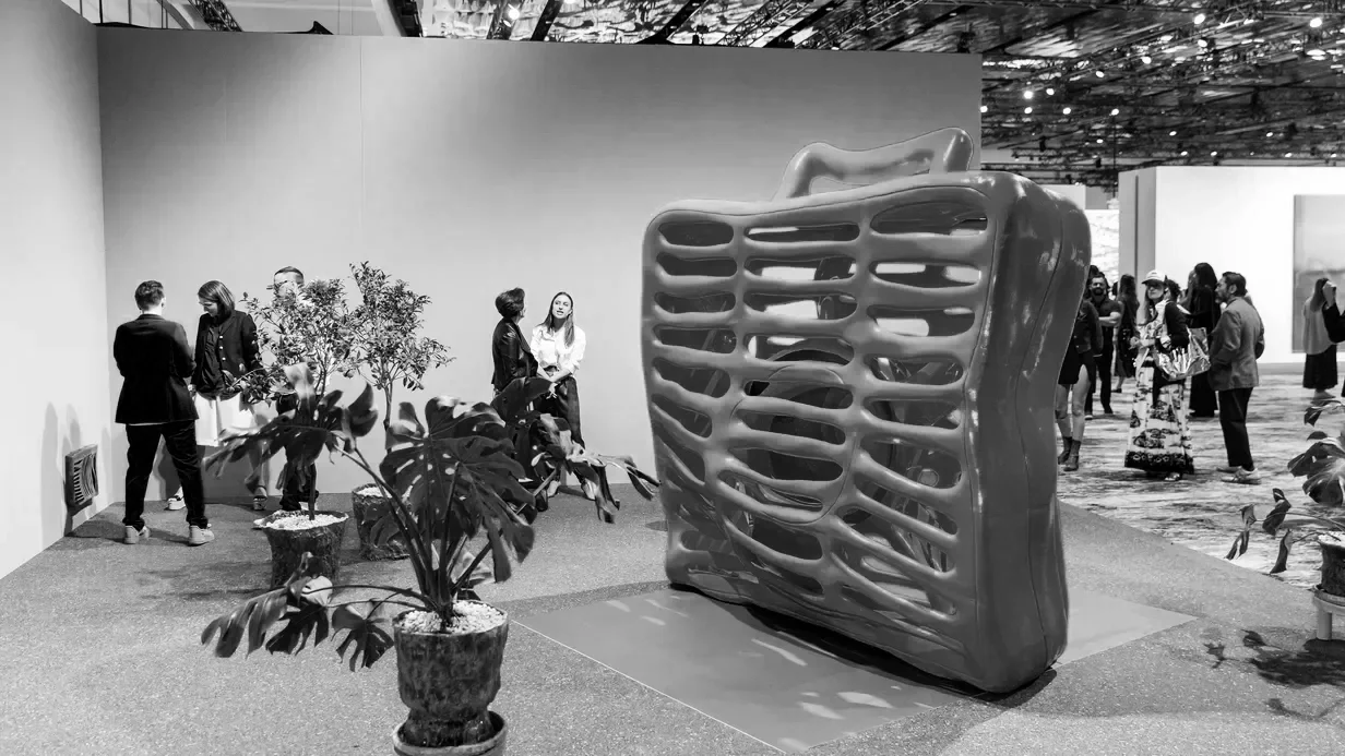 People gathered around a large abstract sculpture resembling a skeletal framework, with potted plants nearby, in an art gallery or exhibition space.