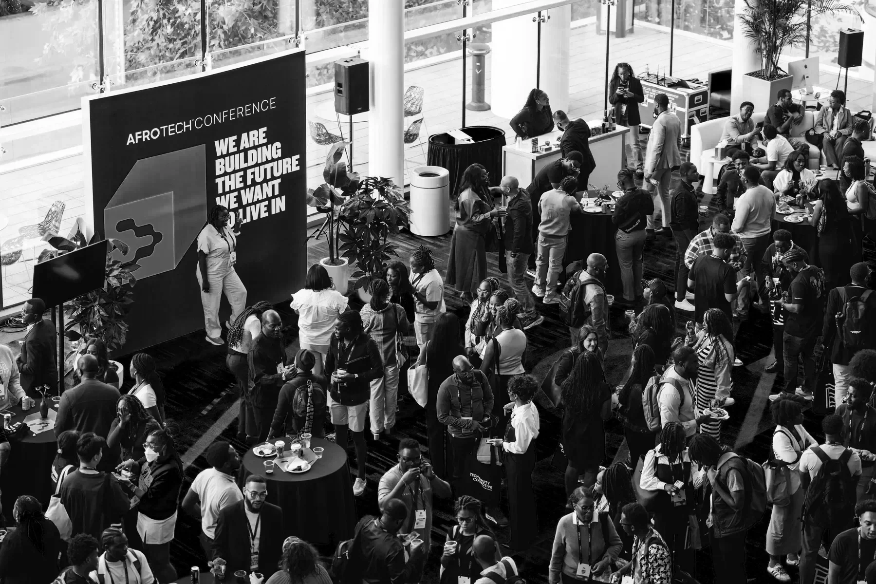 A black and white photo of a busy conference event with many people socializing, some standing and others sitting at tables. There is a large sign that reads 'AFROTECH CONFERENCE' and 'WE ARE BUILDING THE FUTURE WE WANT TO LIVE IN.' The event is held in a spacious, modern venue with large windows allowing natural light, and various booths and seating areas.