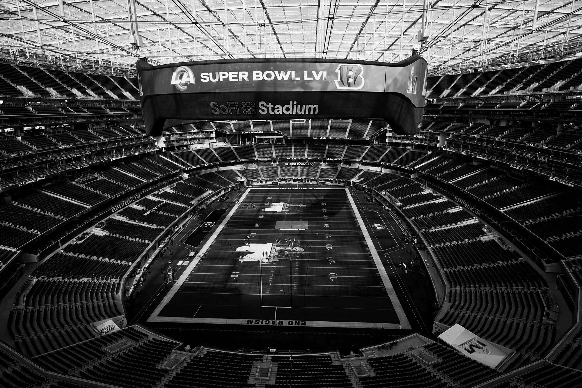 Empty interior of SoFi Stadium with a large digital display showing Super Bowl LVI and NFL logos, and a football field with players practicing.
