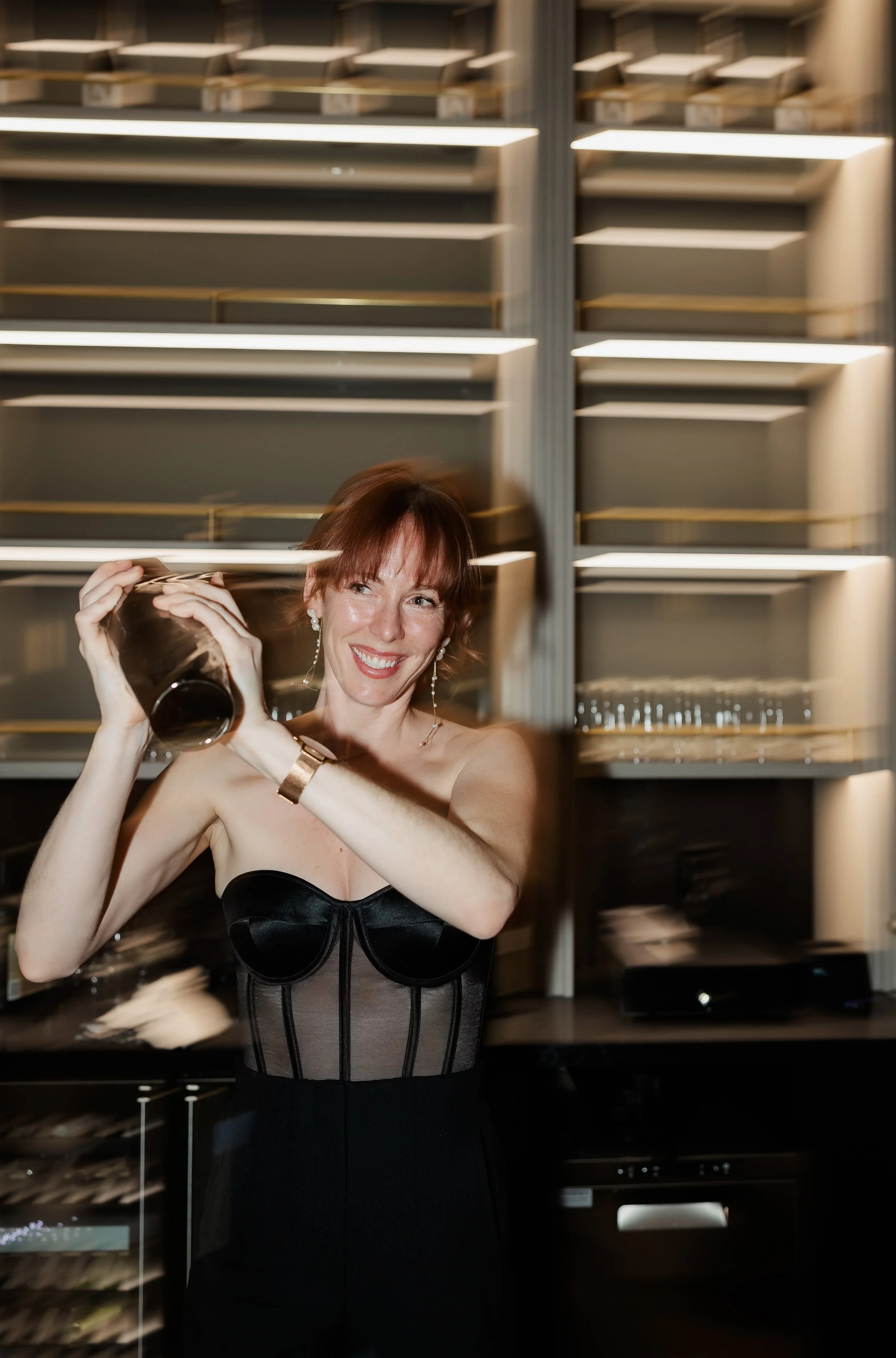 A woman with red hair smiling and holding a shaker in a dimly lit room with shelving and alcohol behind her.