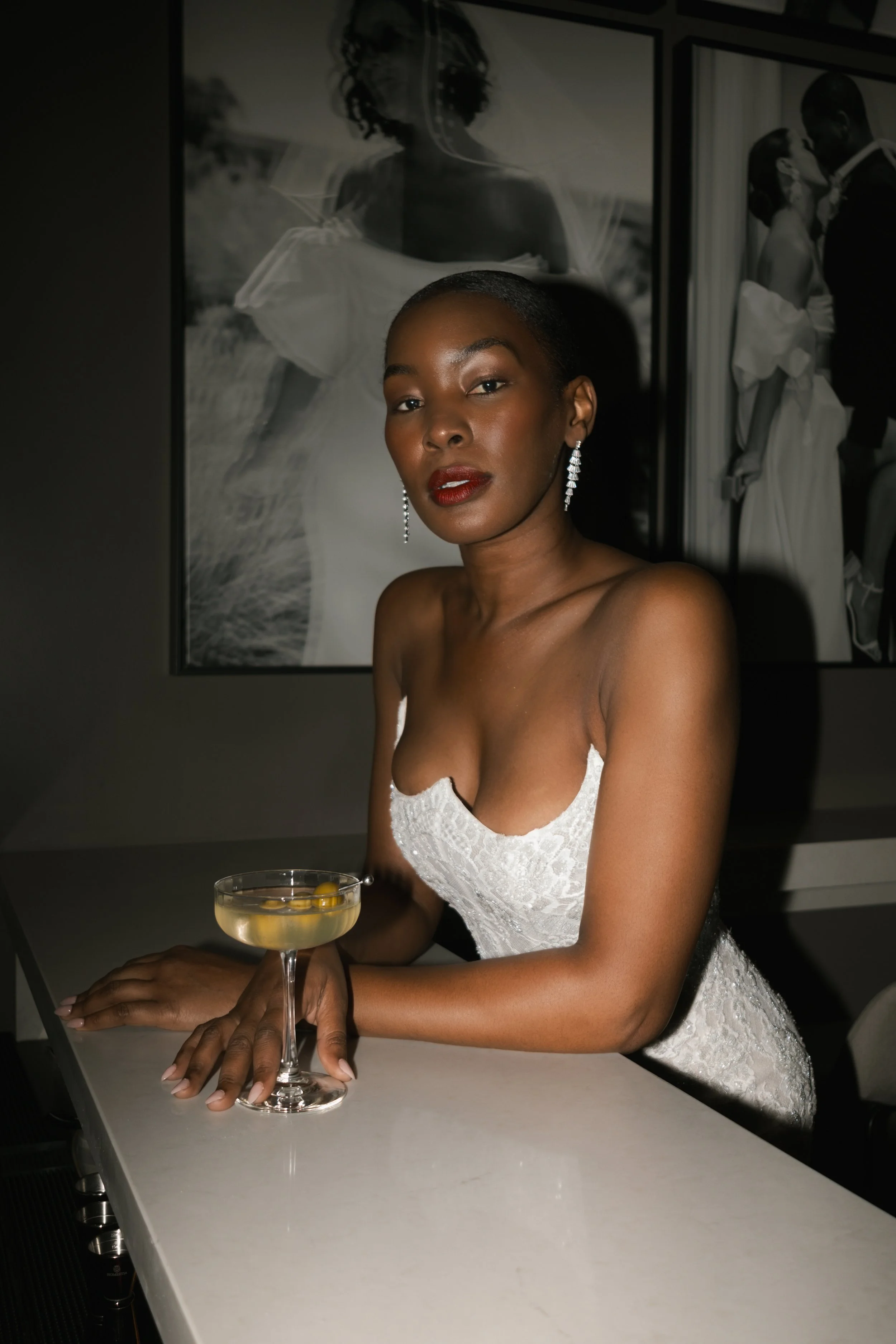 A woman in a white dress and diamond earrings sitting at a bar with a cocktail in a stemmed glass on a white counter, holding the glass with one hand, with black and white wedding photos in the background.
