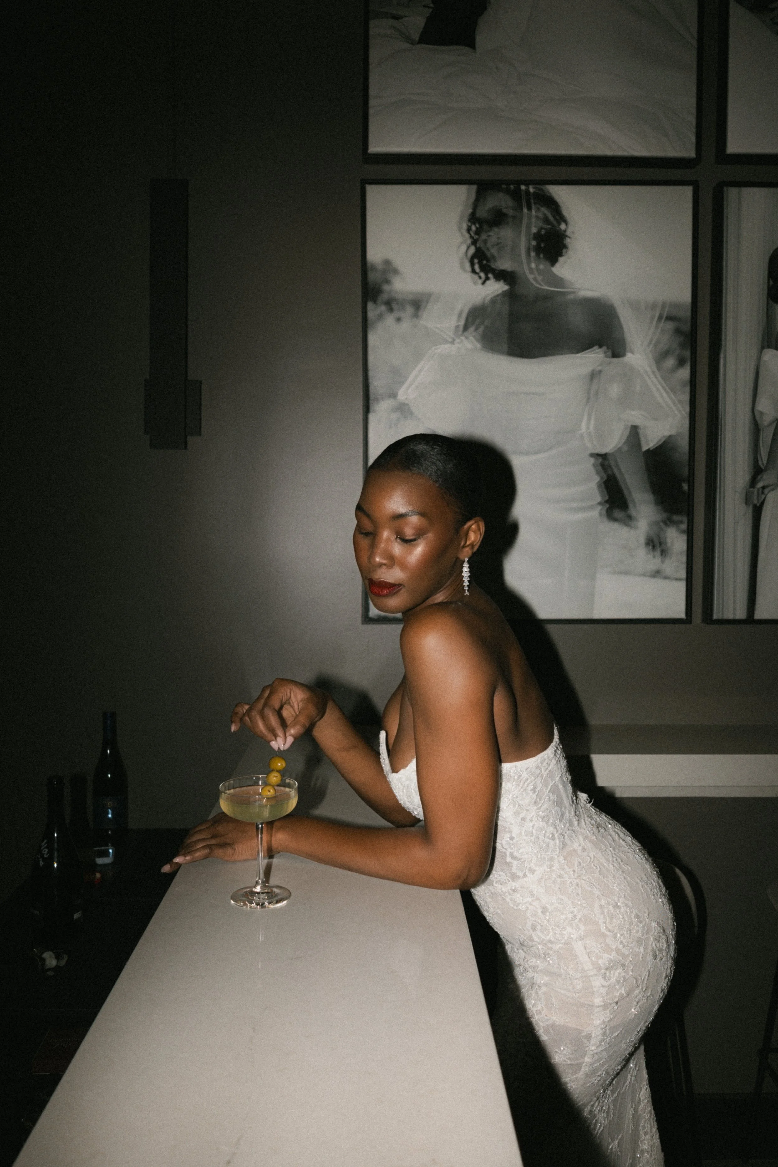 A woman in a white lace off-the-shoulder dress with short hair and earrings is standing at a white bar counter, preparing a cocktail with olives. The background is dark, with a large black-and-white photo of a woman in a dress hanging on the wall.