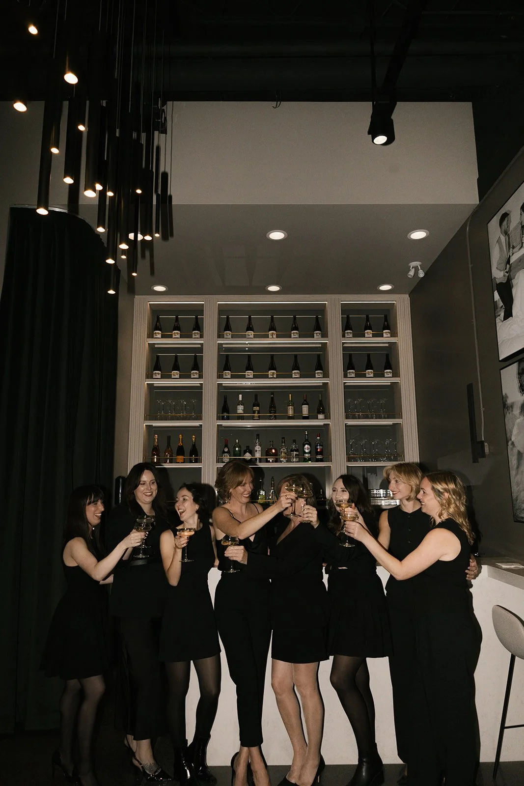 Group of seven women in black dresses enjoying drinks together at a celebration in a speakeasy bar, with a backlit shelf of bottles behind them.