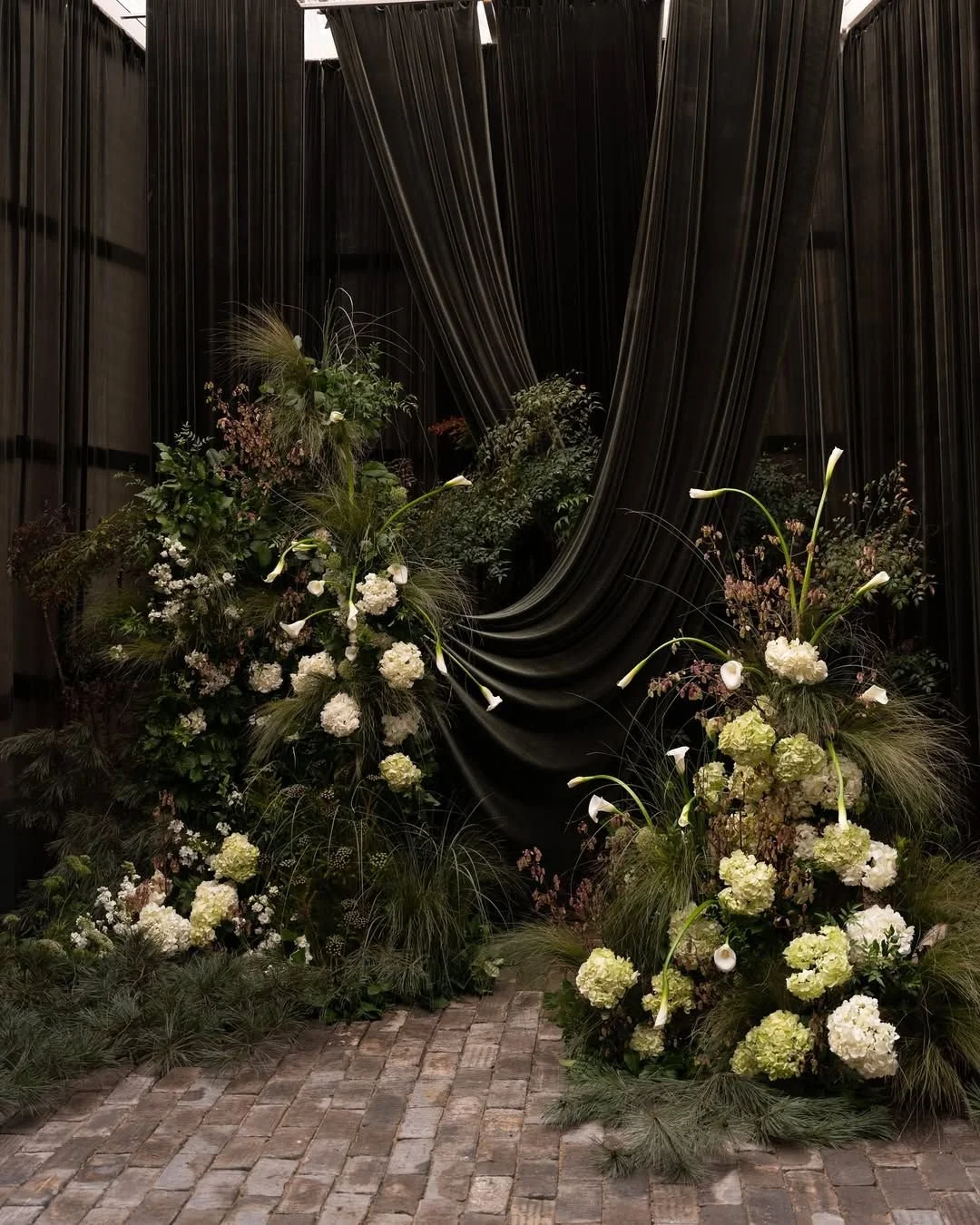 Elegant floral arrangement with white and green flowers, including calla lilies and hydrangeas, placed on a cobblestone floor in front of a green draped fabric backdrop.