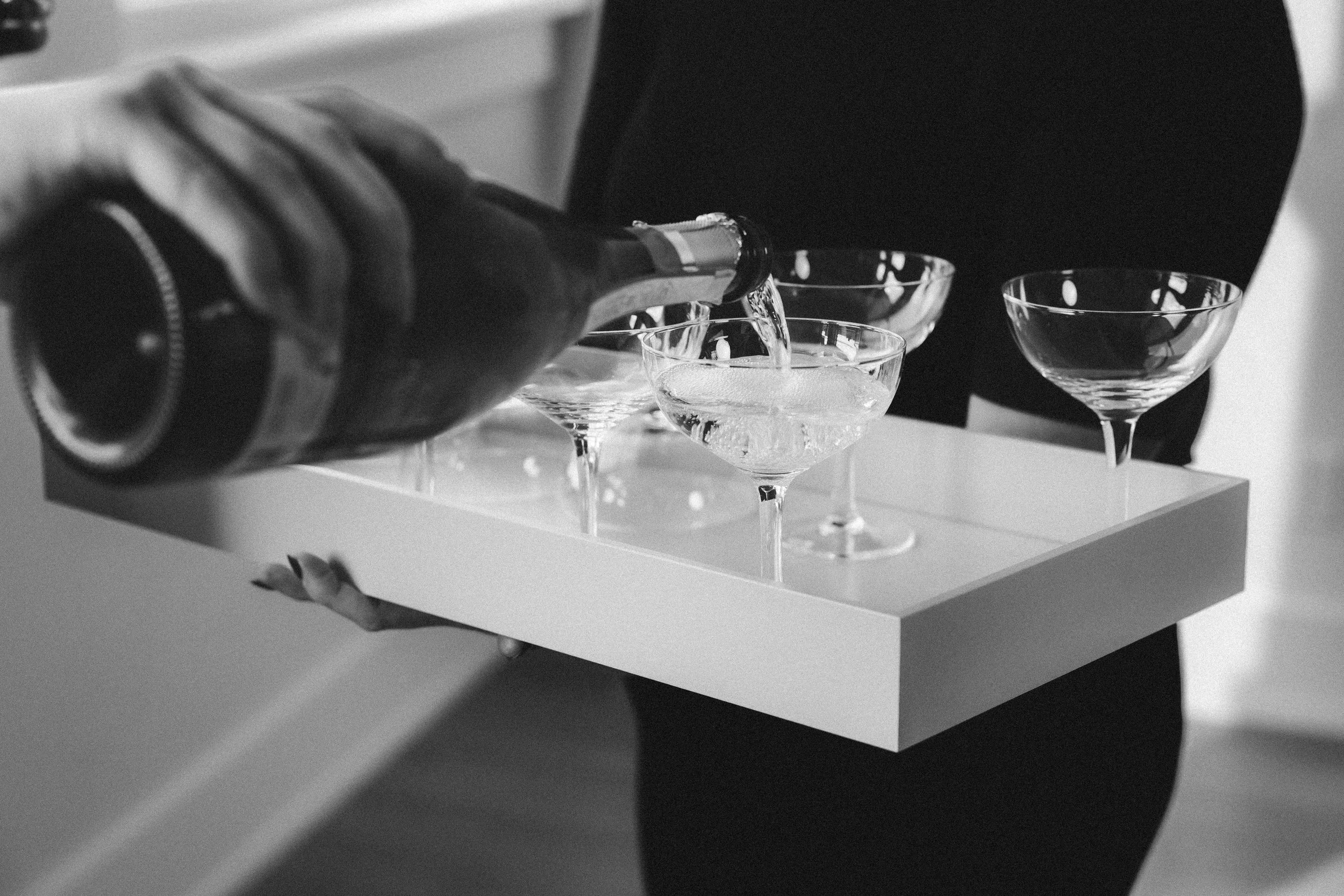 A person pouring a beverage into cocktail glasses arranged on a tray.