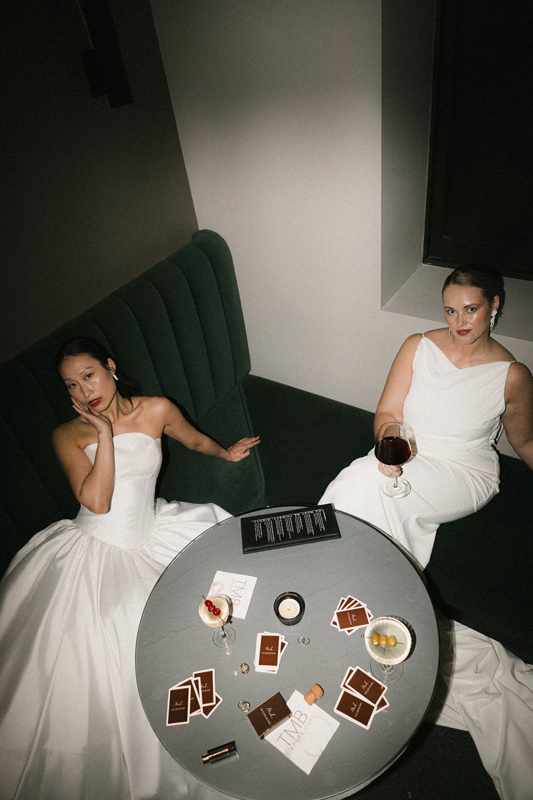 Two women in white dresses sitting on a black booth seat at a round table with drinks, game cards, and a candle, photographed from above in a dimly lit setting.