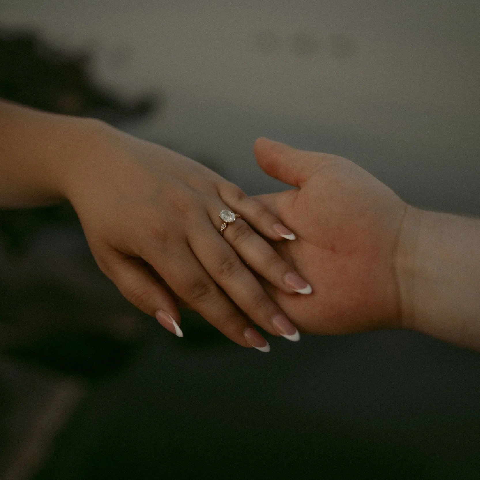 Close-up of two hands gently touching. One hand has a large engagement ring on the ring finger, with manicured nails.