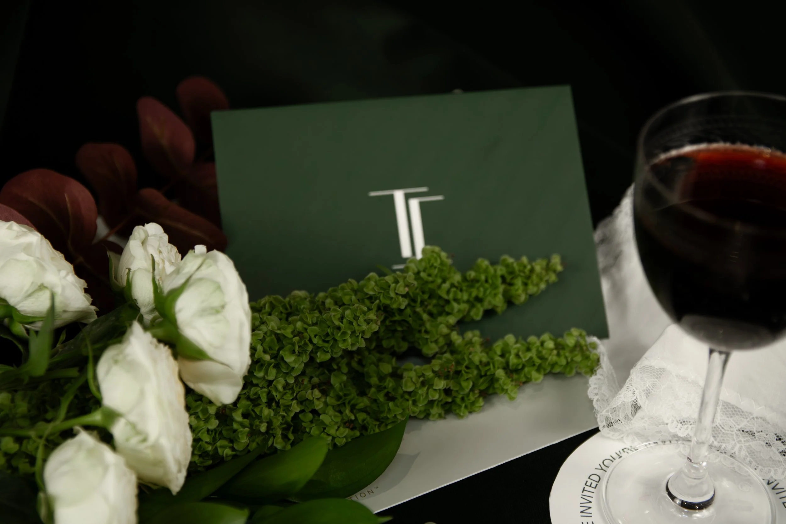Close-up of a floral arrangement with white roses, green hydrangeas, and red leaves, accompanied by a glass of red wine, a green card with a white logo, and a white lace cloth.