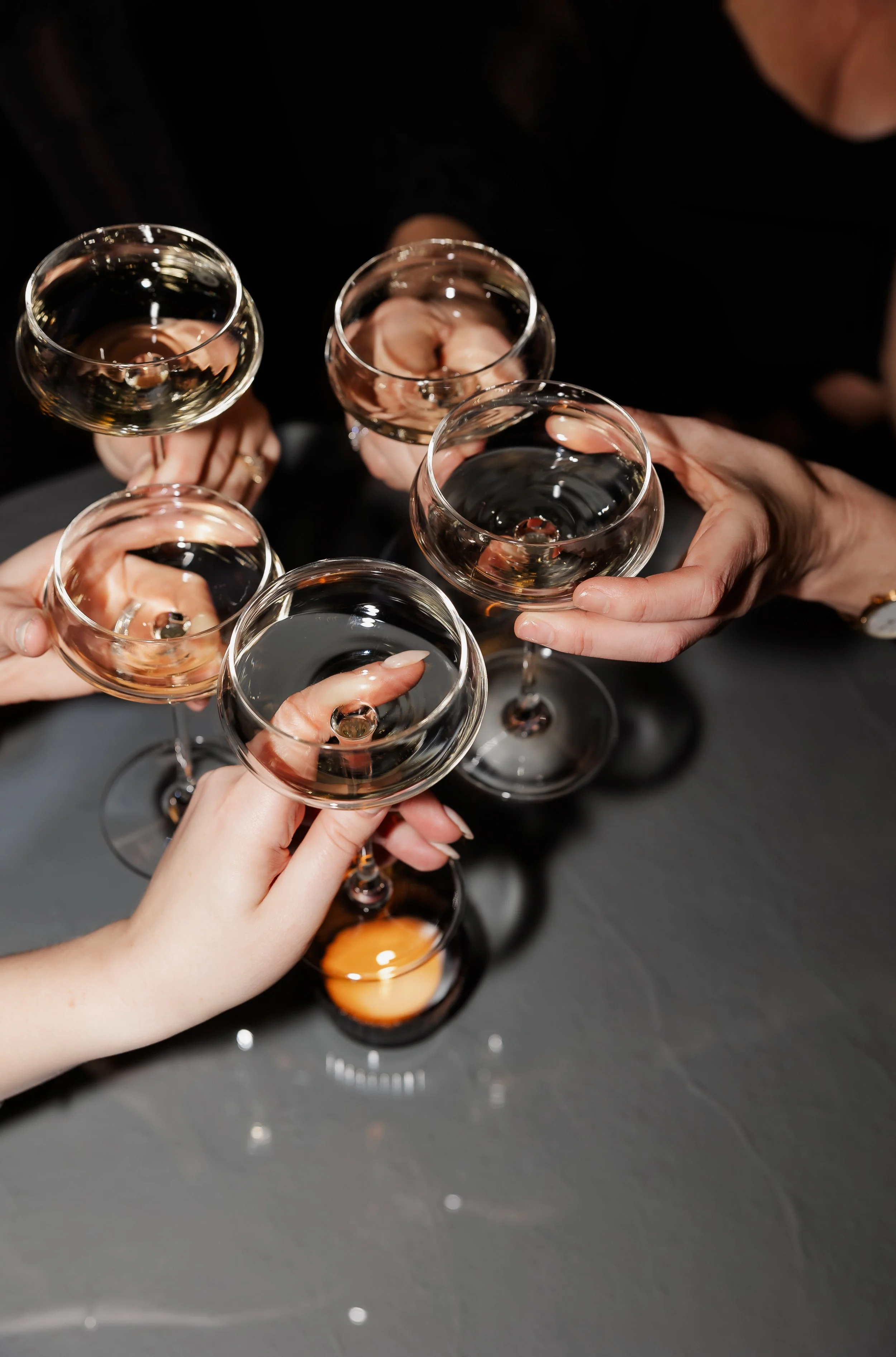Multiple hands holding wine glasses filled with white wine, raised together in a toast against a dark background.