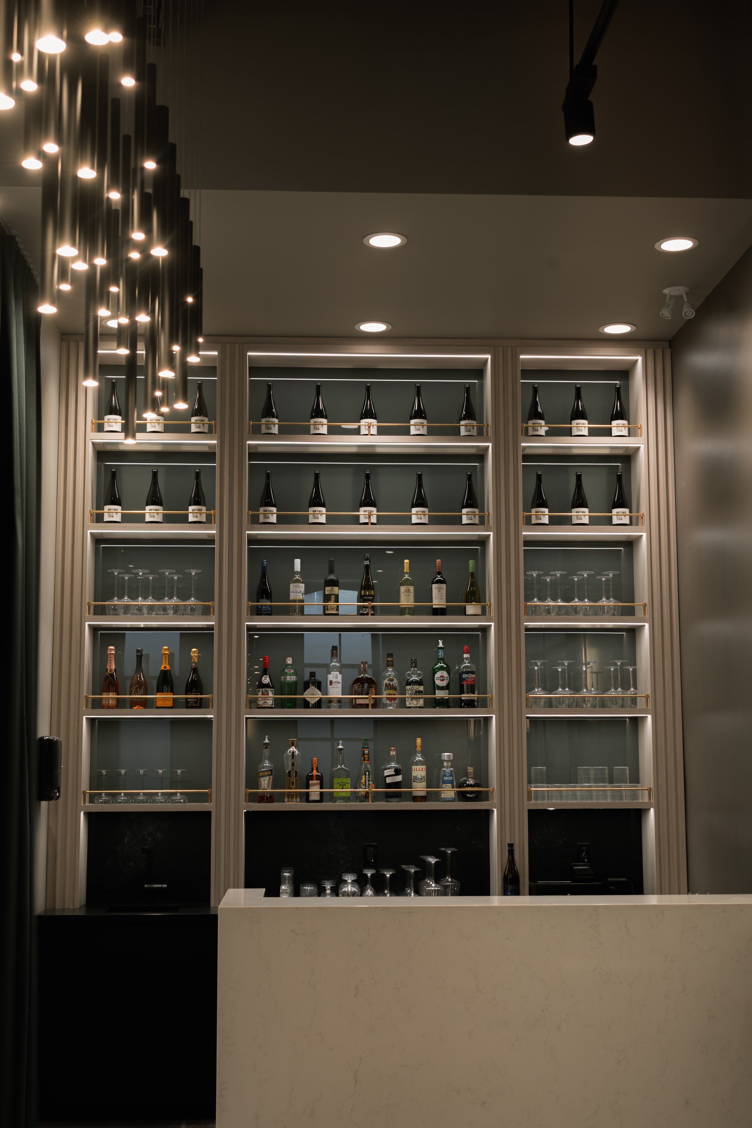 Bar counter with shelves of liquor bottles and wine bottles, glassware on shelves, modern lighting, and a marble counter.
