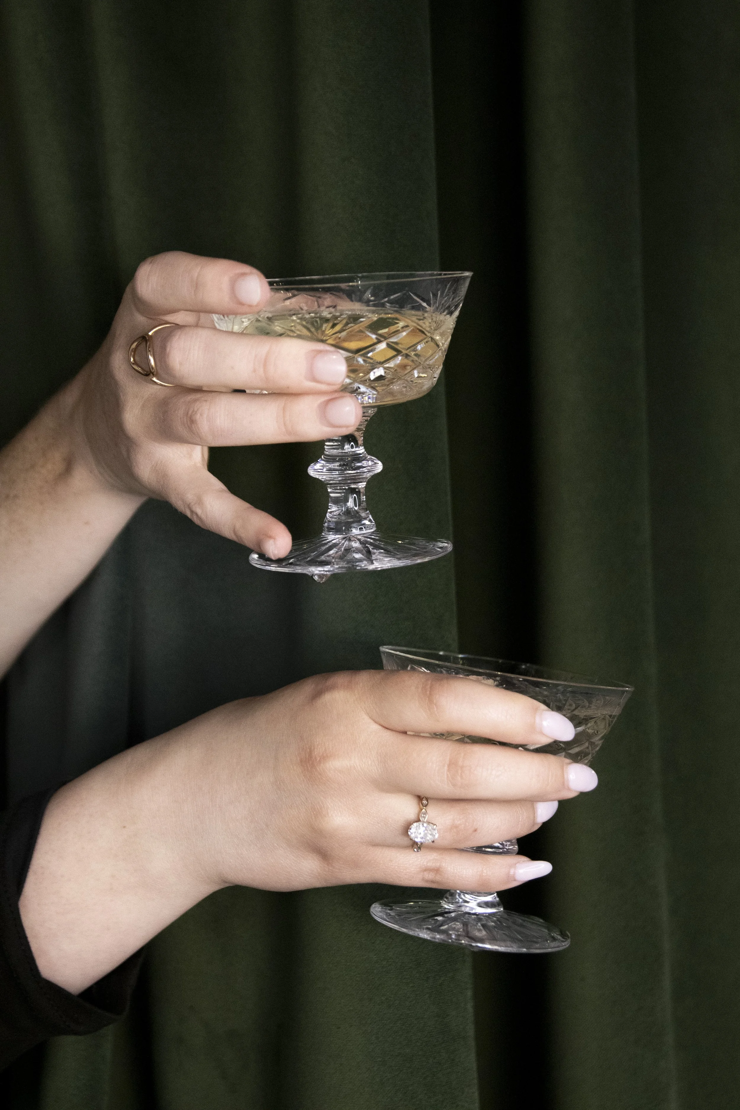Two hands holding vintage glass coupe glasses filled with champagne, against a dark green curtain background.
