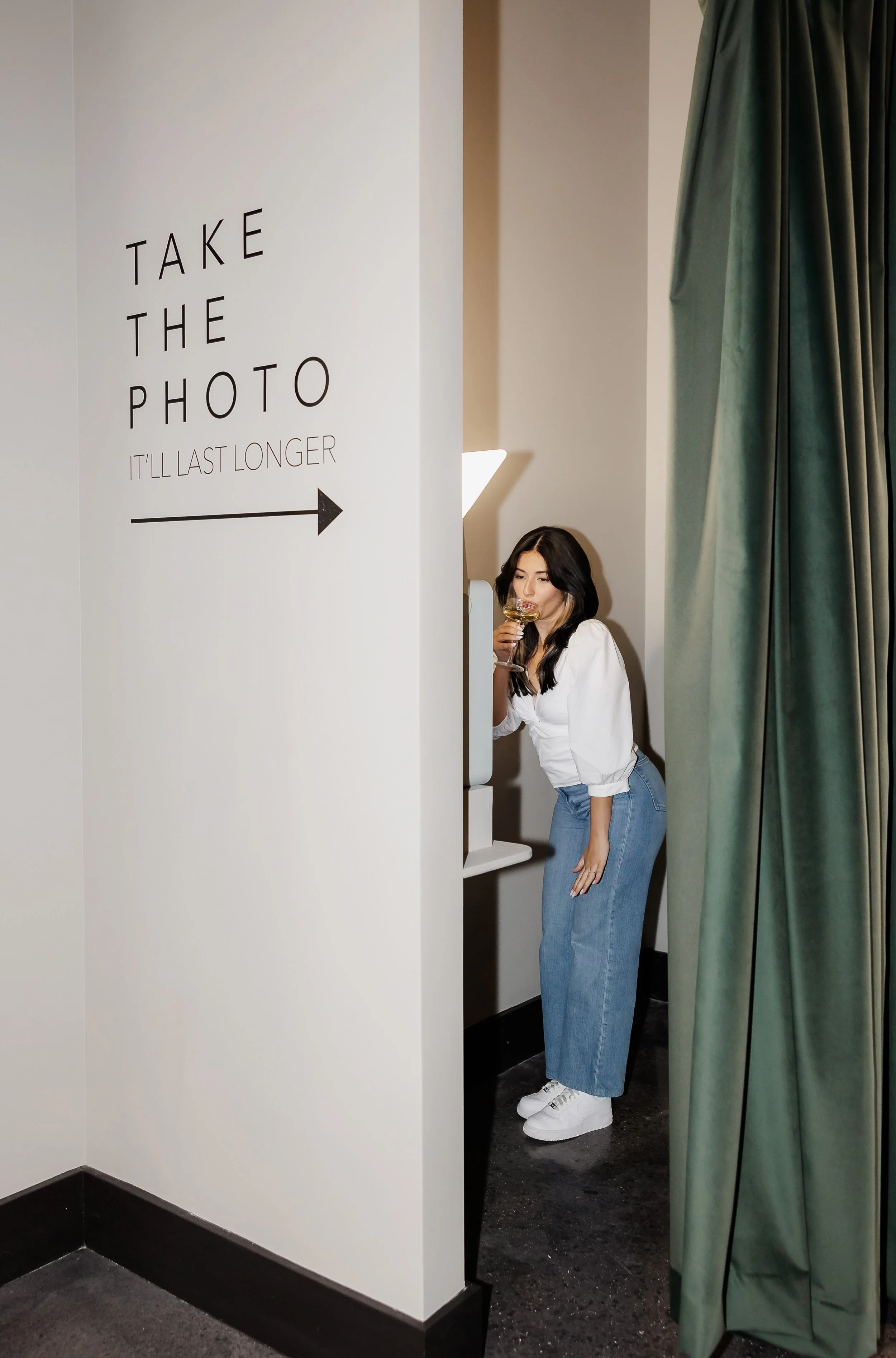 A young woman with long black hair, wearing a white long-sleeved top, blue jeans, and white sneakers, is standing behind a curtain and looking at a photo booth with a drink in her hand. There is a sign on the wall that says 'Take the photo, it'll las
