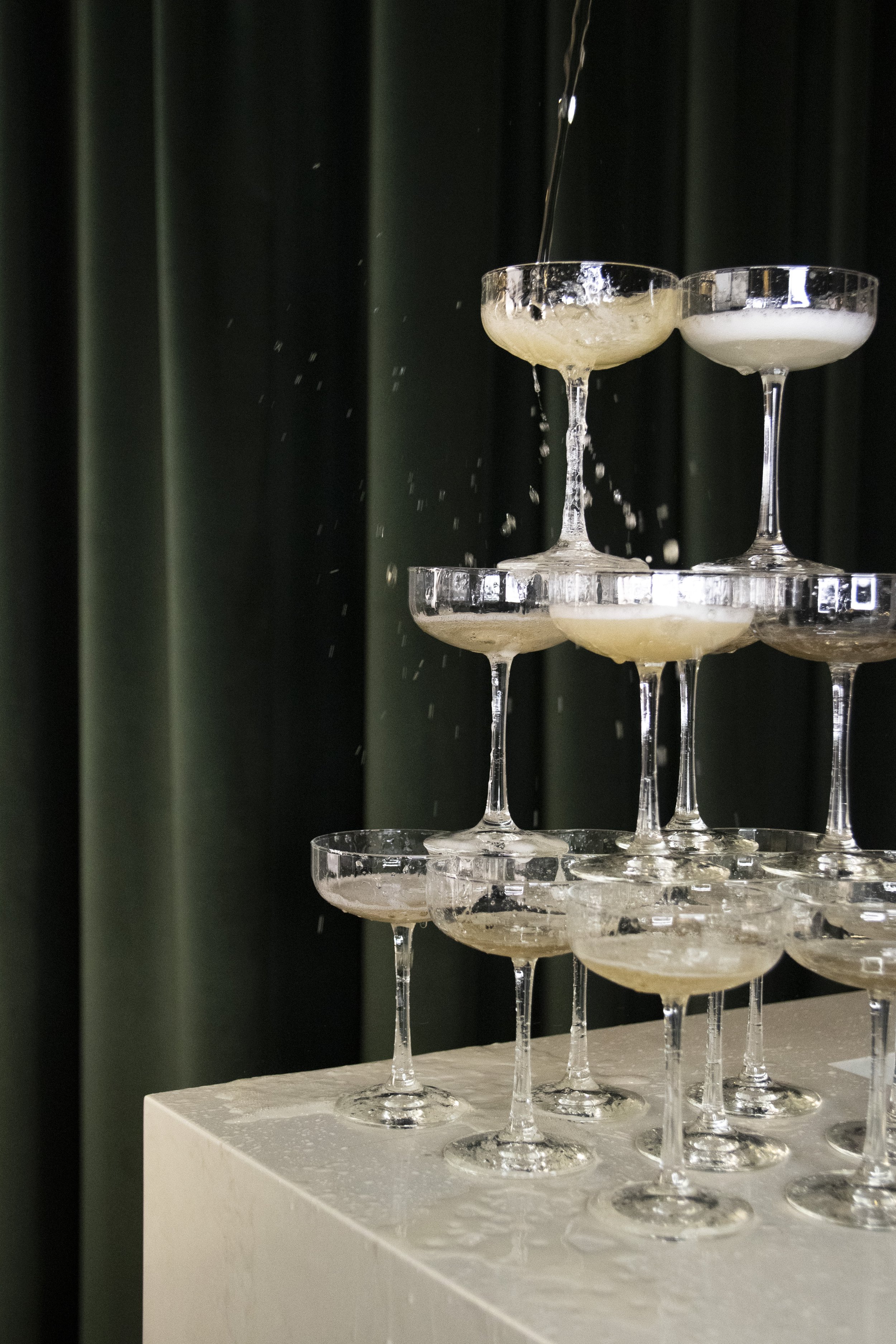 A champagne tower with multiple tiers of empty champagne glasses, some with foam, set against a dark curtain background.