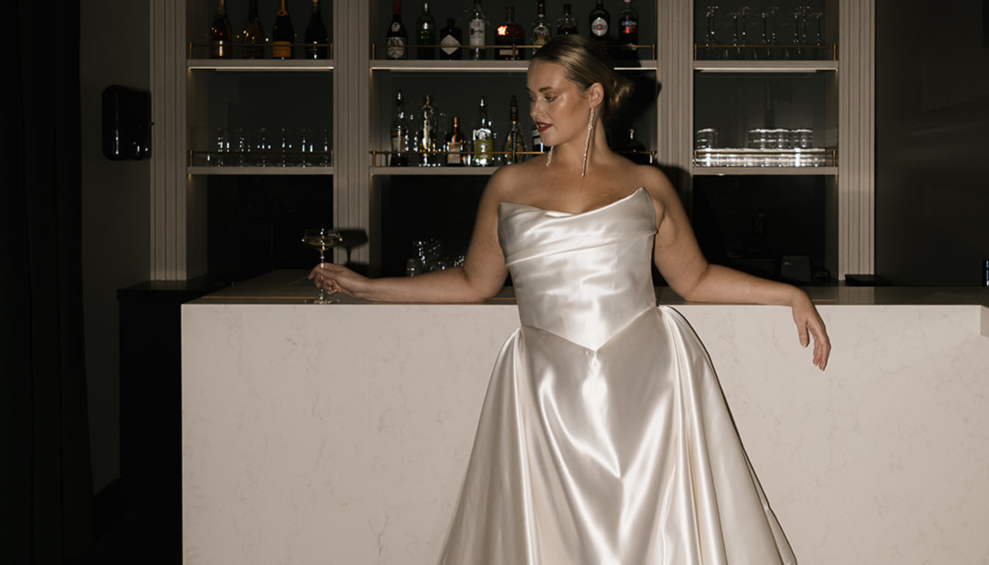 A woman in a satin wedding gown standing at a bar, holding a glass of champagne.