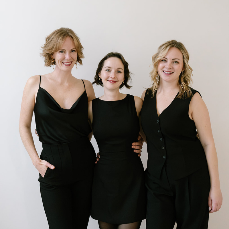 Three women in black outfits smiling and posing together against a white background.