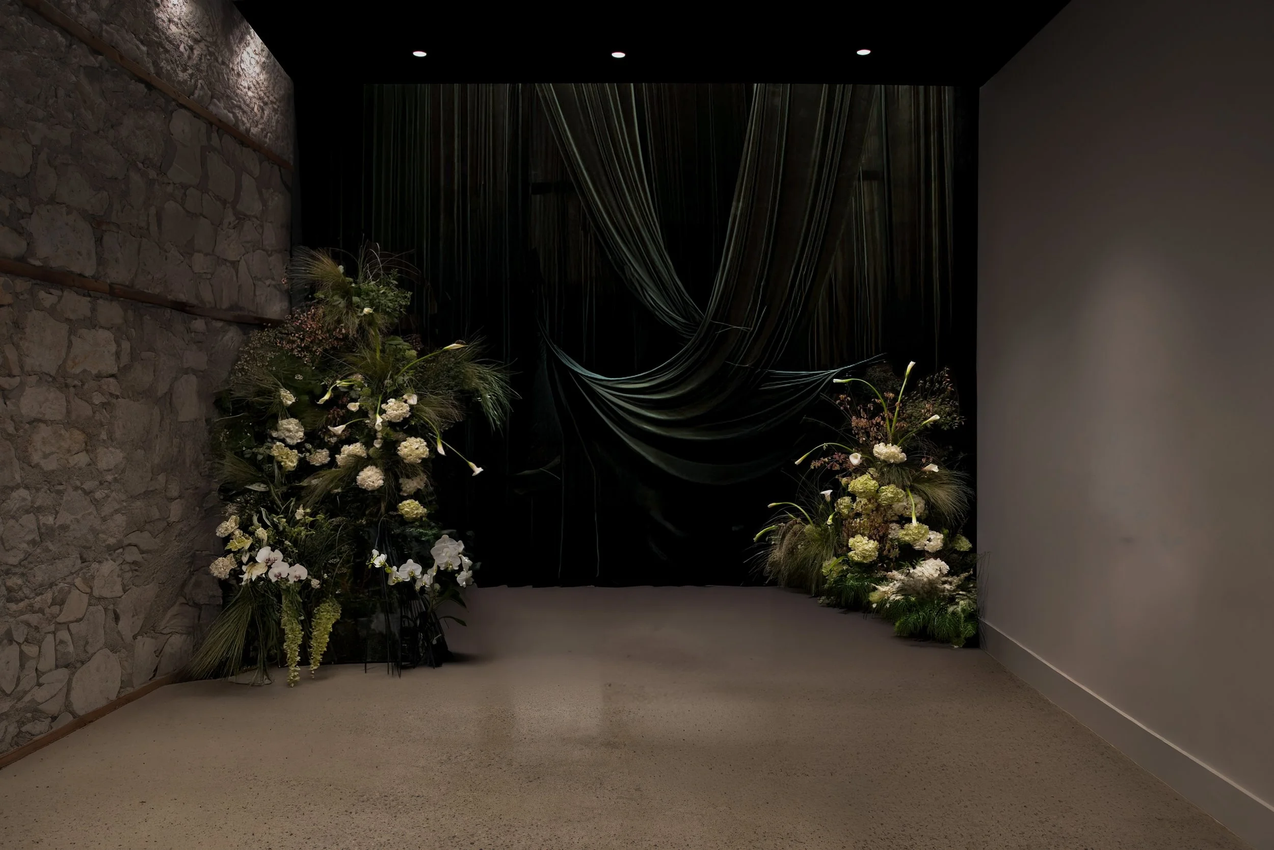 THE INVITE by TMB, decorated with a green fabric backdrop draped with floral arrangements of white and green flowers on either side, set against a stone wall on the left and a plain wall on the right.