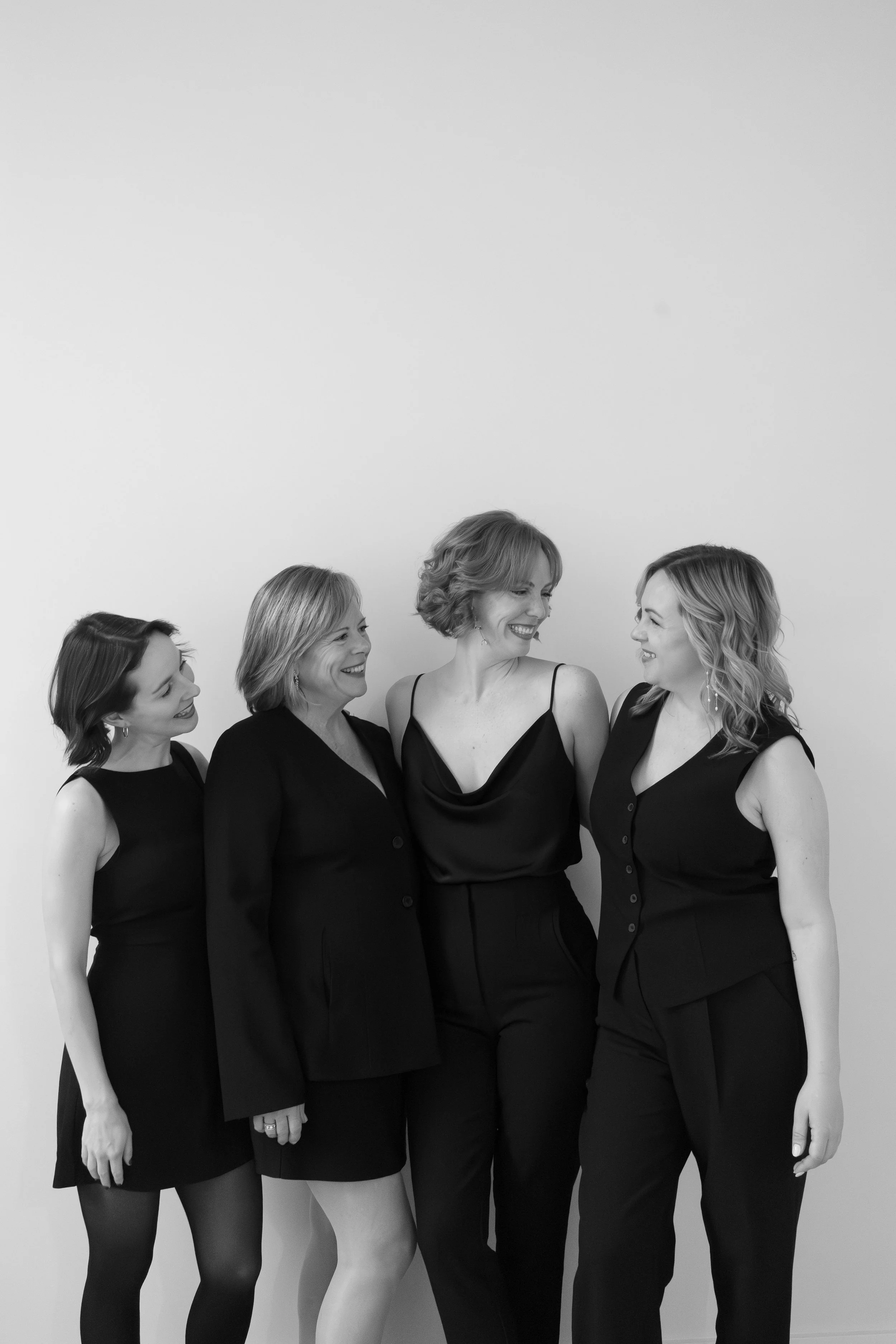 Five women in black dresses standing together and smiling, looking at each other against a plain background.